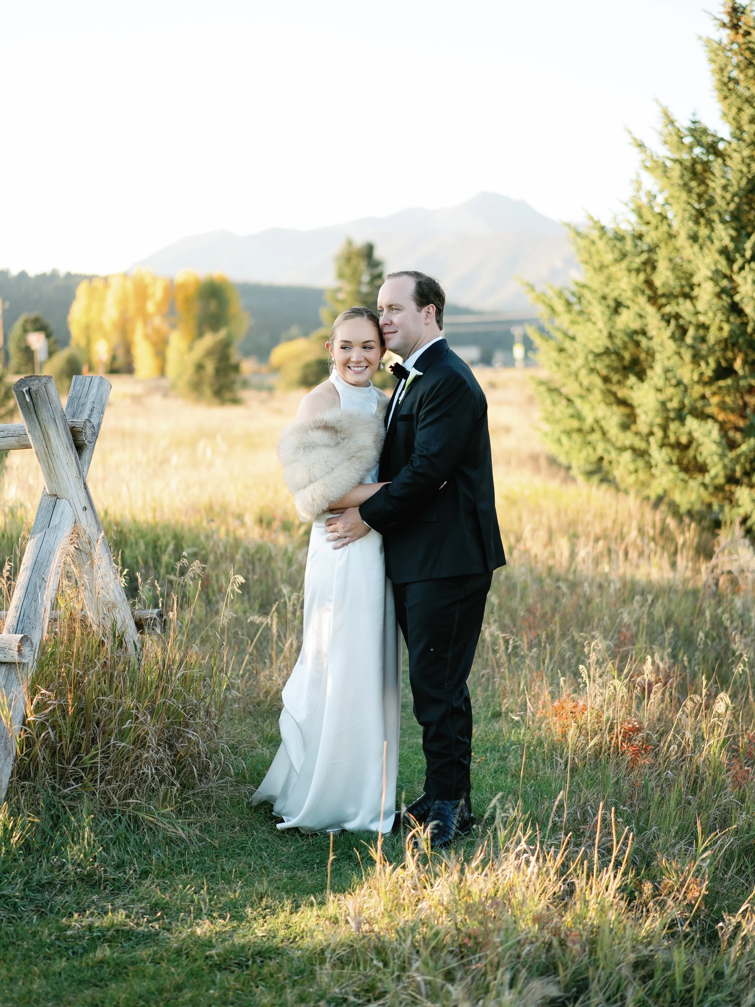 A bride and groom standing together outdoors in a field during sunset, smiling, with mountains and trees in the background.