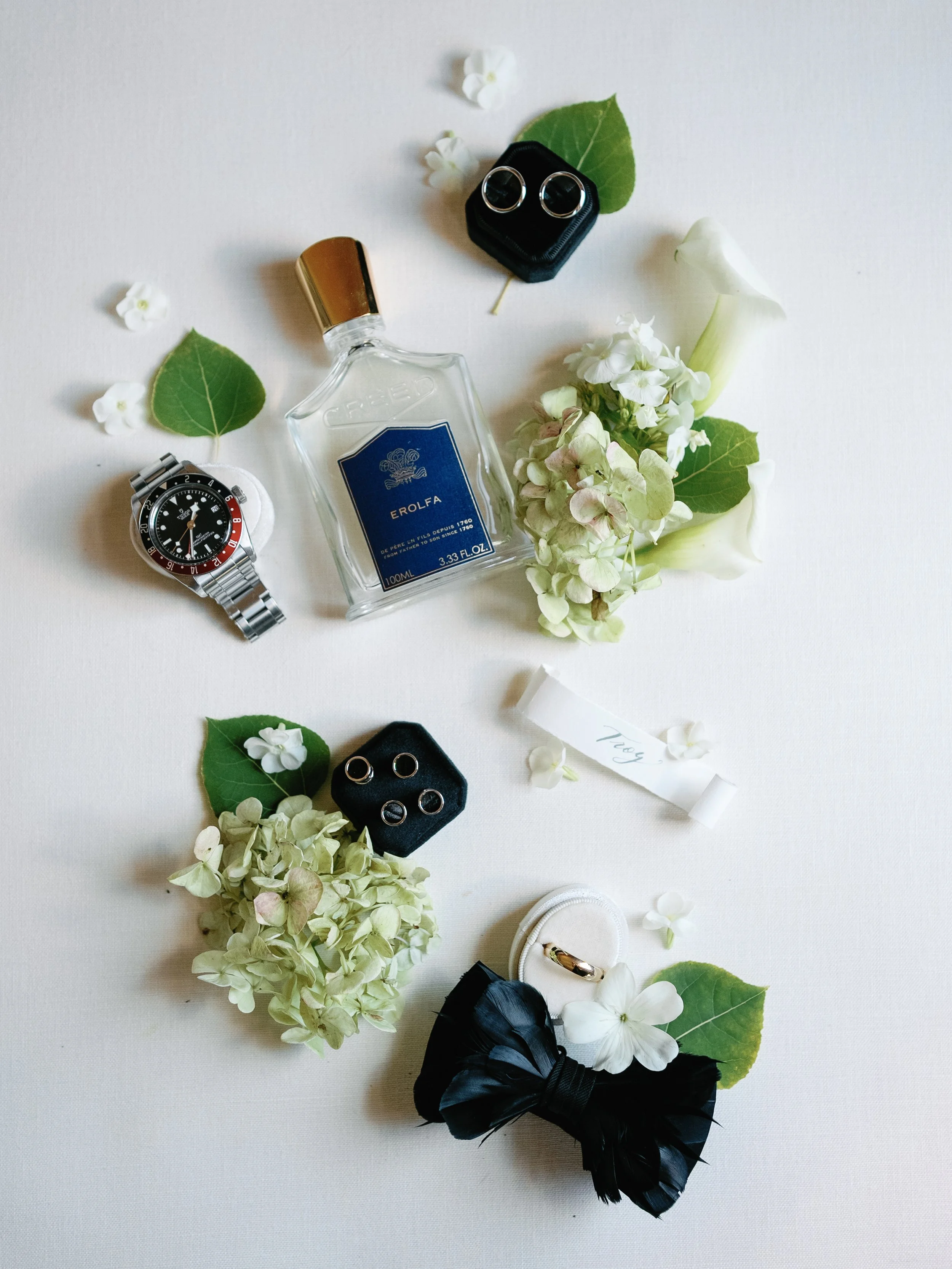 Watch, ring, perfume bottle, jewelry rings, earrings, flower boutonniere, flower petals, black hair bow, white ring box, small white roll of paper with writing.