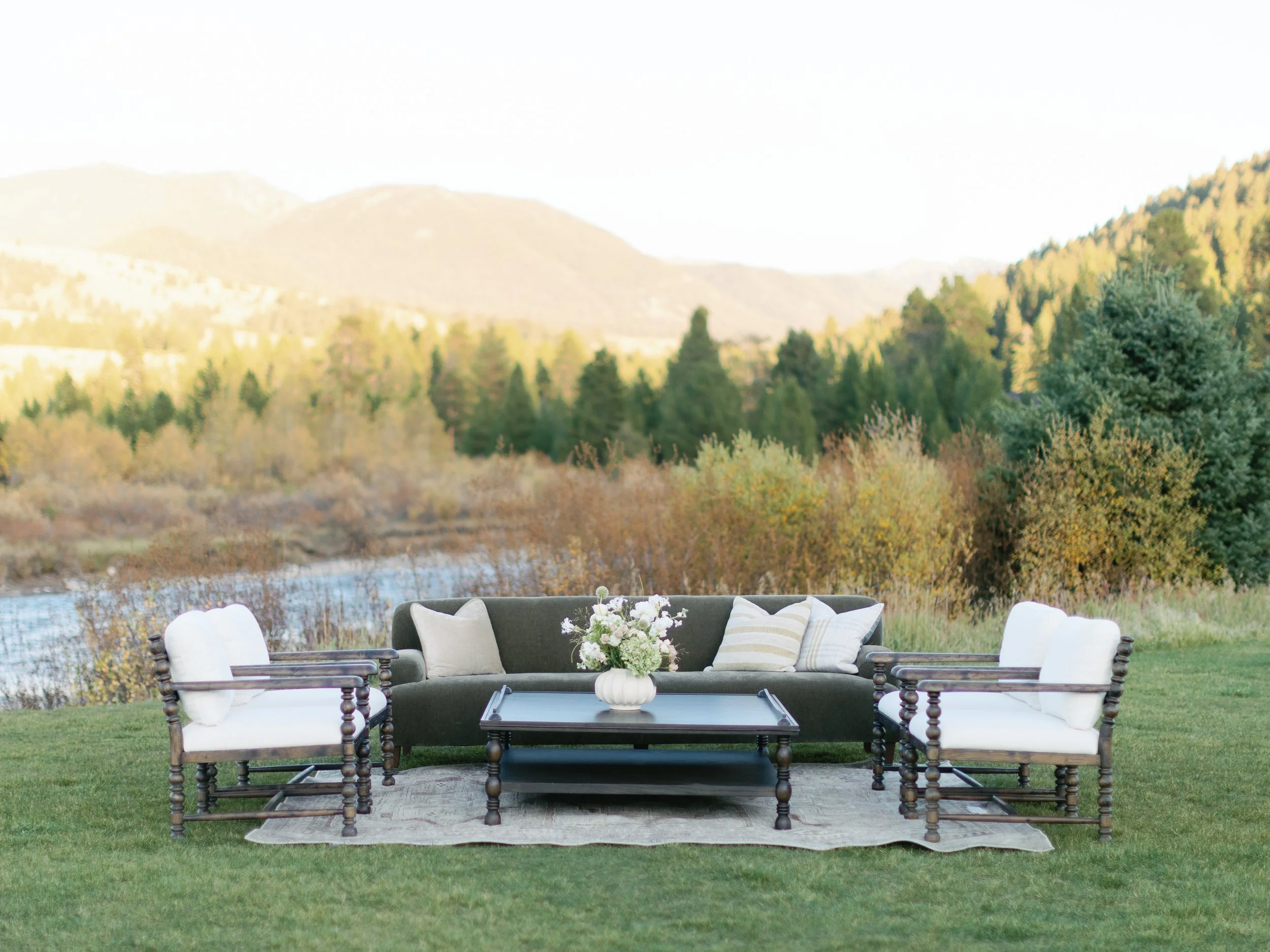Outdoor patio furniture set with a dark sofa, white cushioned chairs, a coffee table with a flower vase, and pillows, set on a grassy area with a river, trees, and mountains in the background under bright daylight.