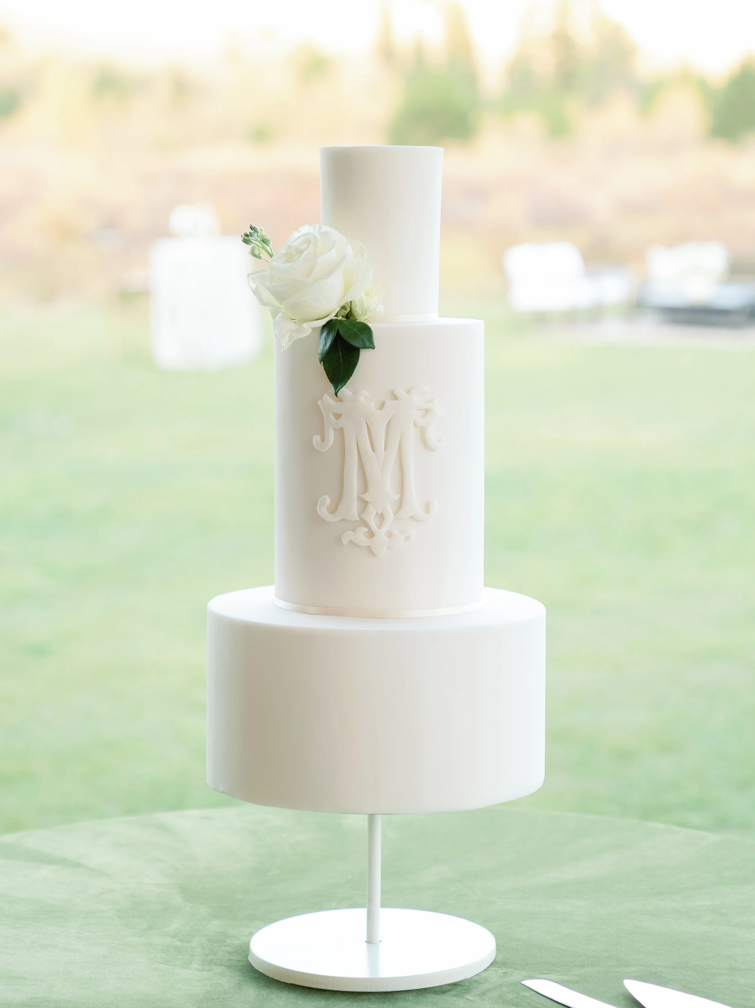 Elegant white wedding cake with three tiers, decorated with a white rose and green leaves, and a monogram letter 'M' on the middle tier, set against a blurred outdoor background.