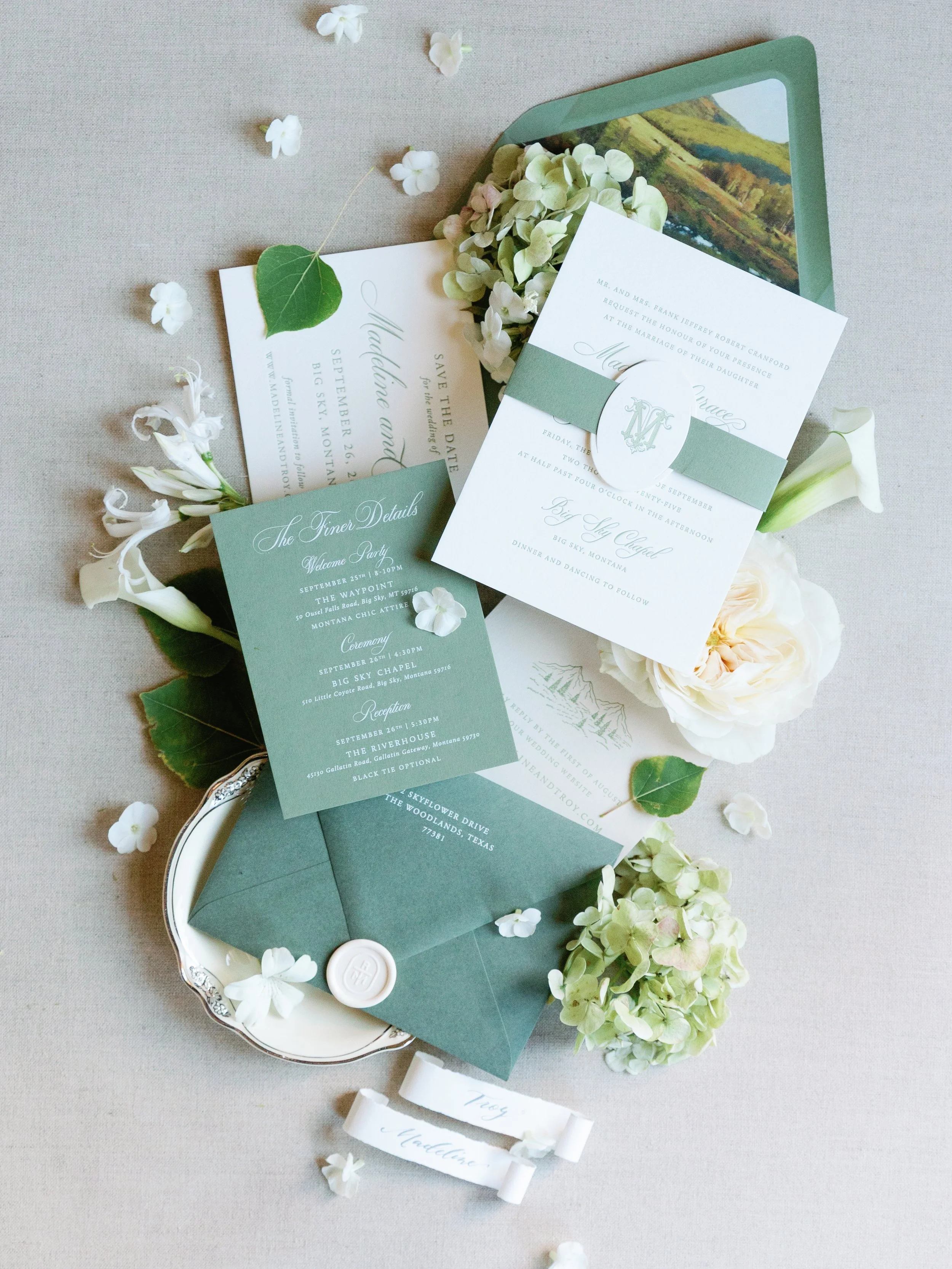 Wedding invitation suite with white and green stationery, surrounded by white flowers and green leaves on a neutral background.