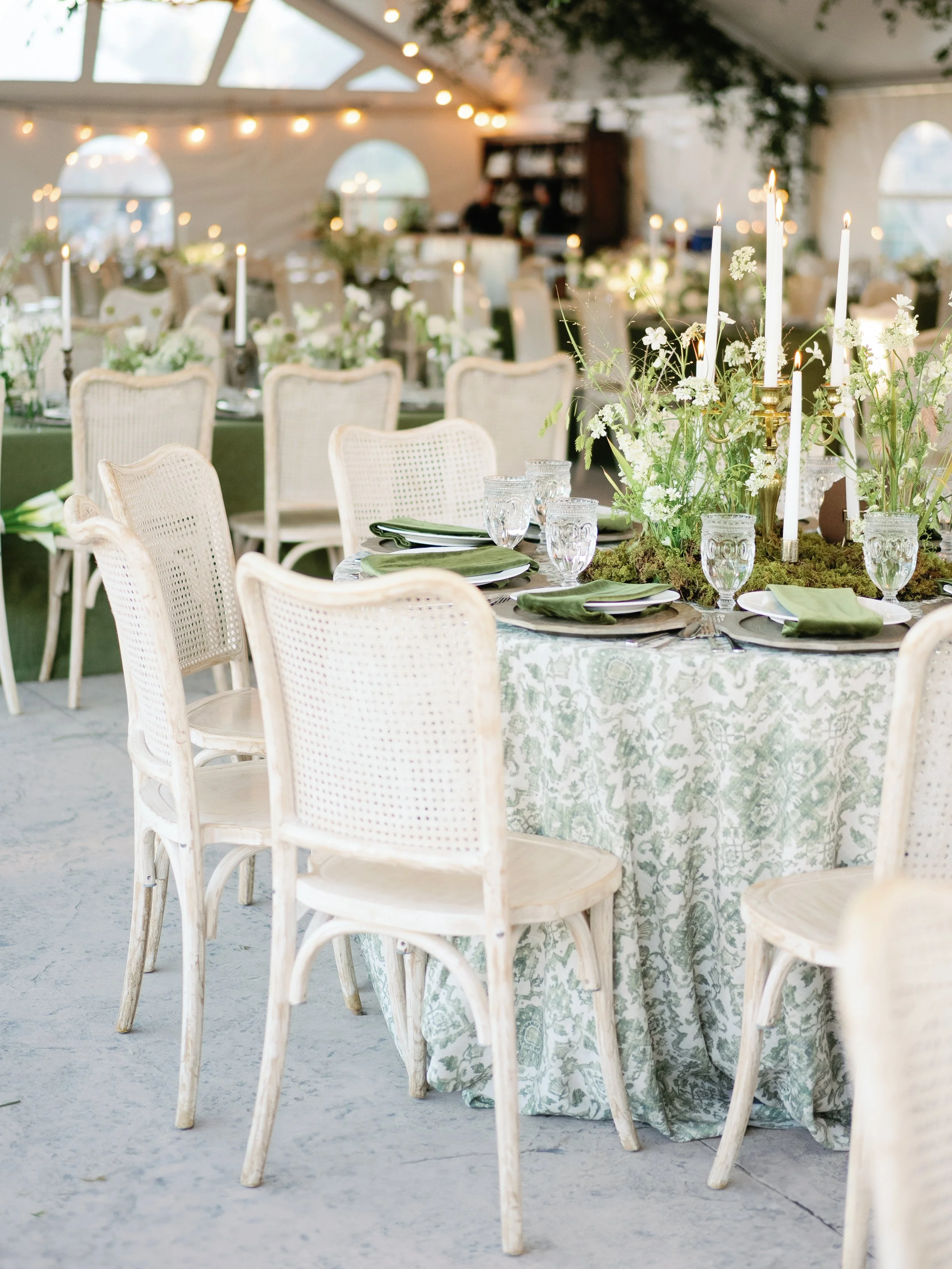 Elegant dining setup inside a tent with round tables, white chairs, green tablecloths, and floral centerpieces with candles, fairy lights, and greenery.