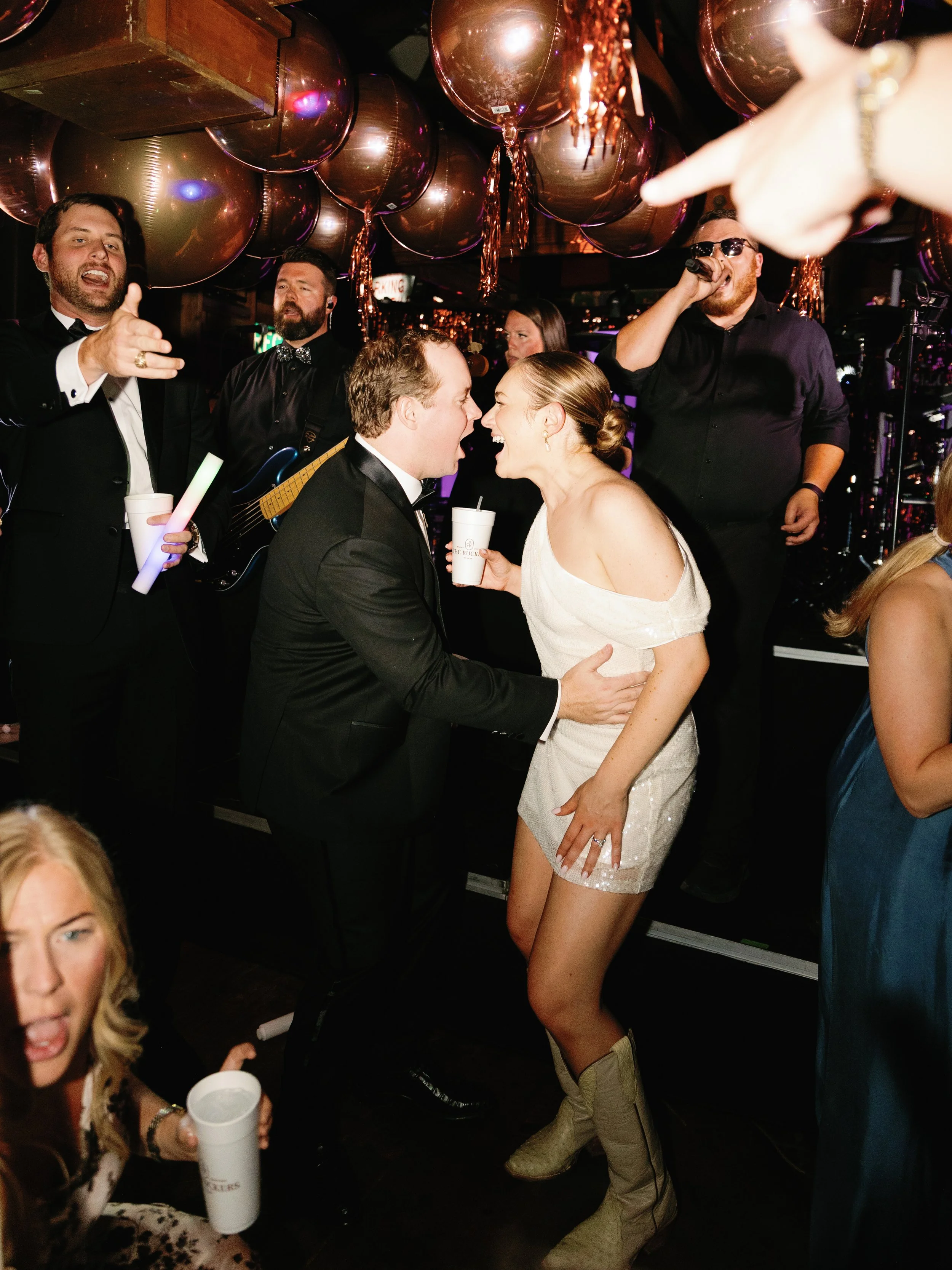 A lively party scene with a couple in formal wear kissing in the center, surrounded by people dancing and celebrating, with balloons and festive decorations overhead.