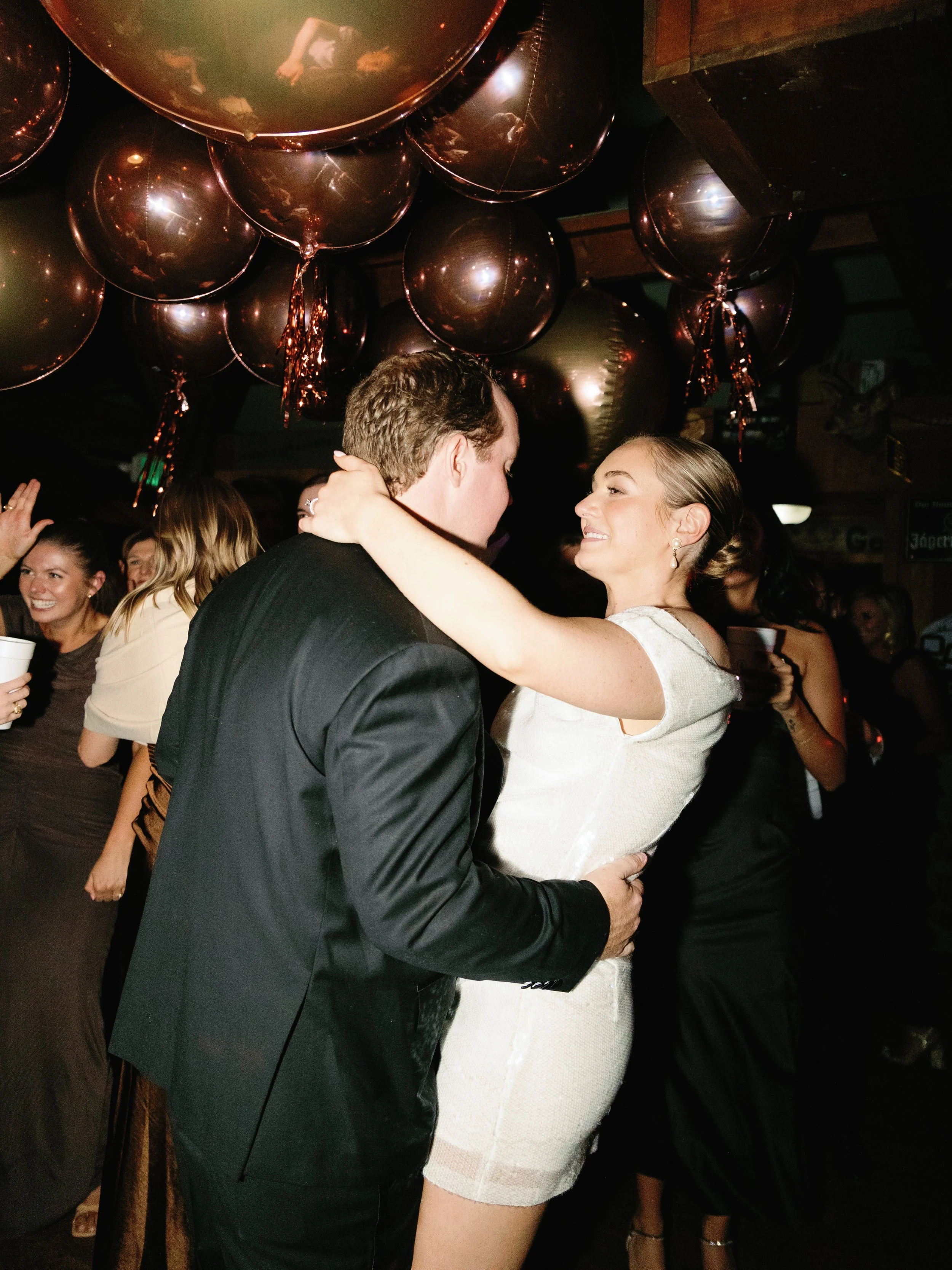 A couple dancing closely at a party or celebration, surrounded by other smiling partygoers, with balloons hanging from the ceiling.