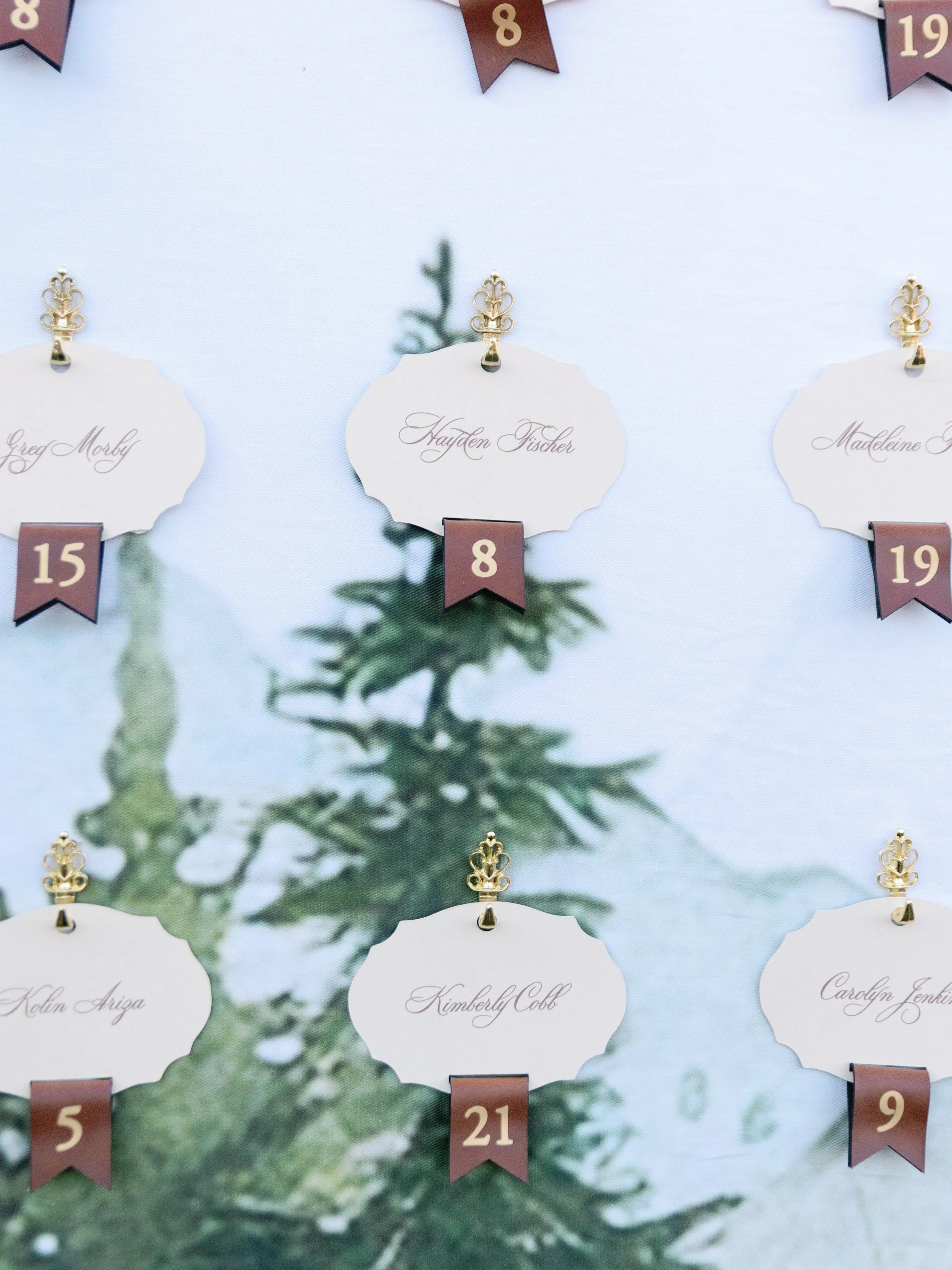 A wedding seating chart with small white cards hanging from gold clips, each with a guest's name and a small banner with a table number. The cards are arranged on a backdrop with a pine tree silhouette.