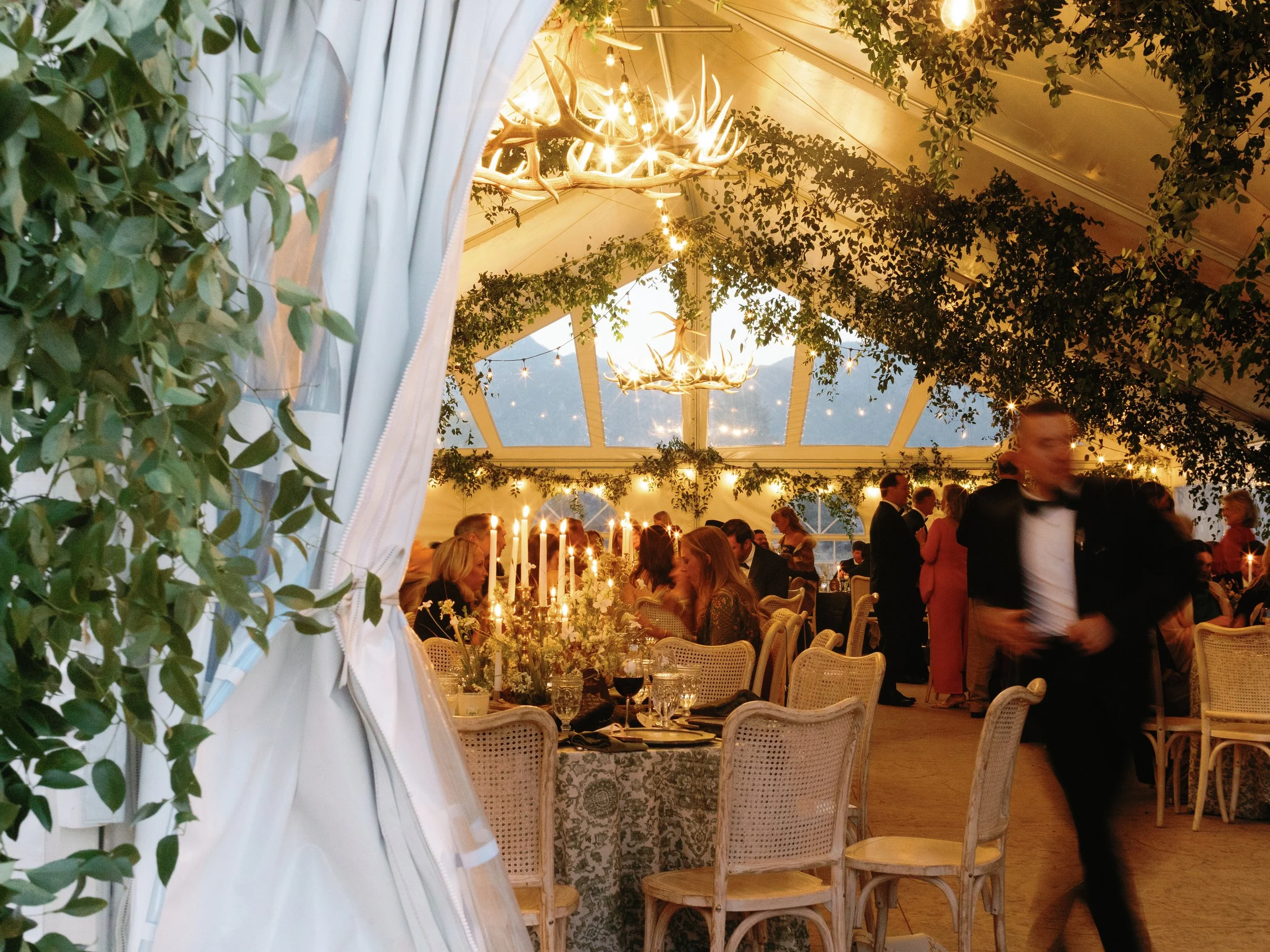 Interior of a decorated wedding reception tent with hanging antler chandeliers, string lights, and greenery. Guests are seated at tables, and a waitstaff in a tuxedo is walking.