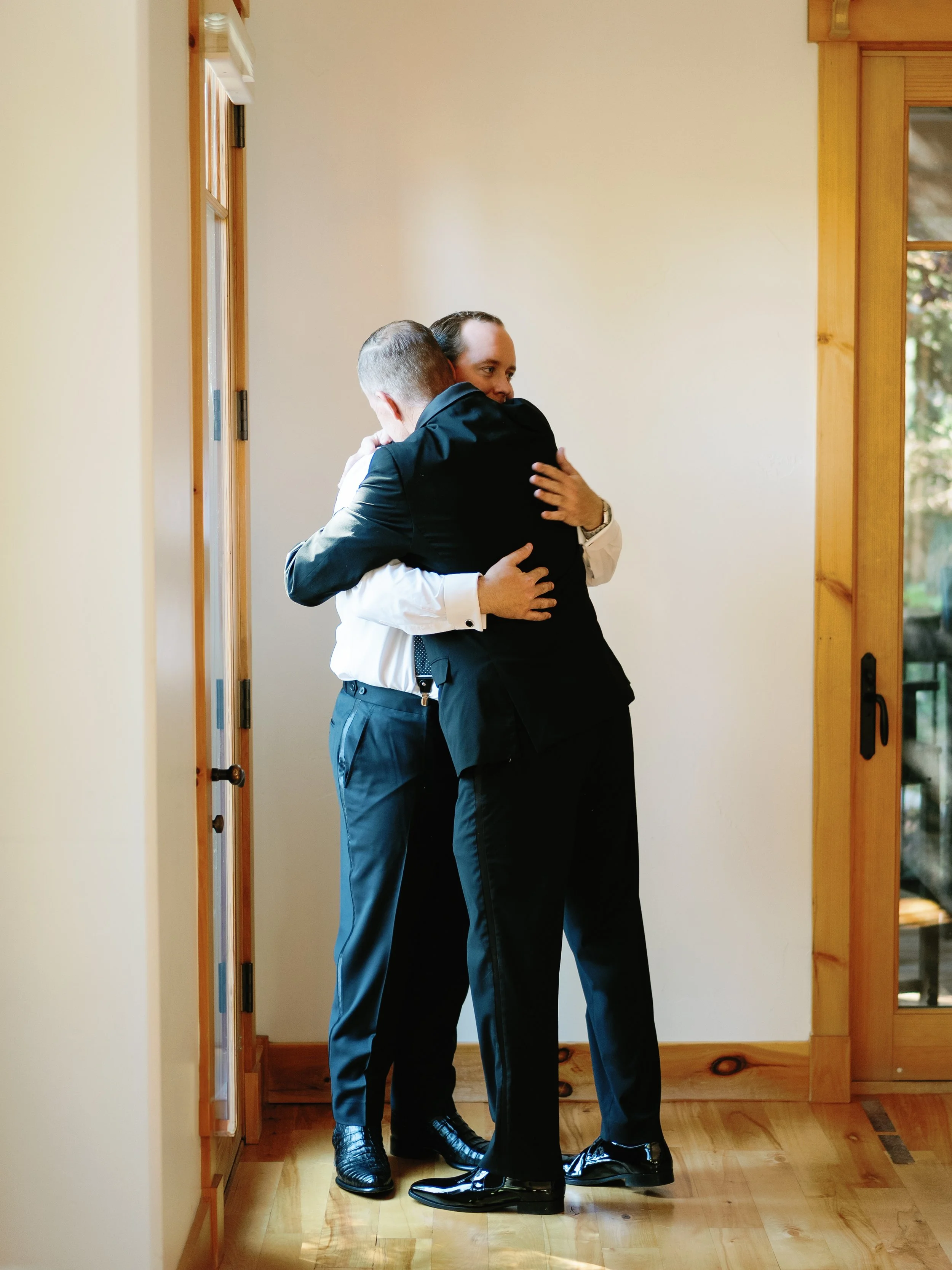 Three men in formal attire embracing each other in a hug inside a room with wooden floors and walls, near a glass-paneled door with a view of trees outside.