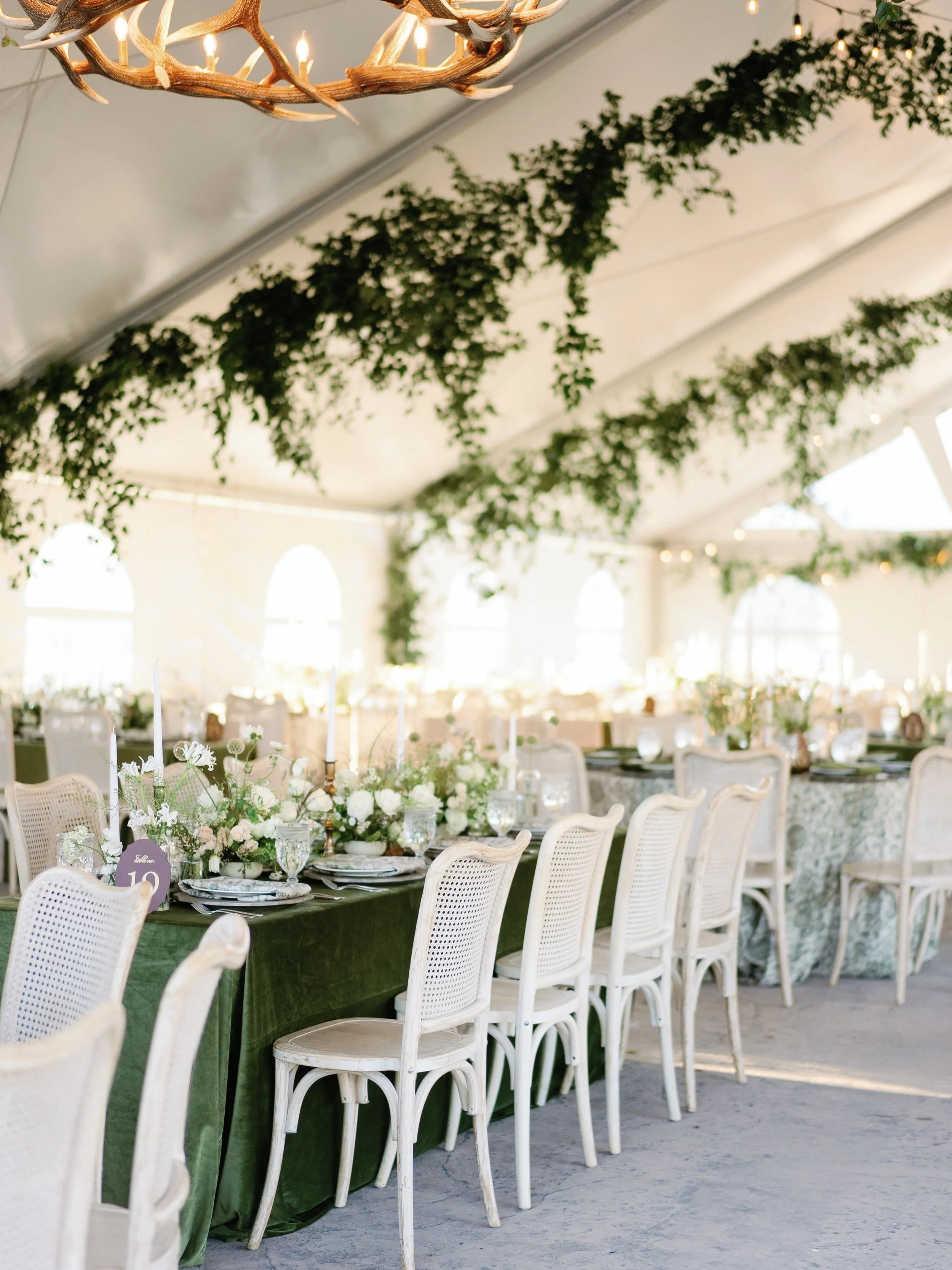 Elegant indoor event setup with long tables covered in dark green tablecloths, decorated with white floral centerpieces, candlesticks, and fine china. White chairs are arranged around the tables under a white ceiling adorned with hanging greenery and