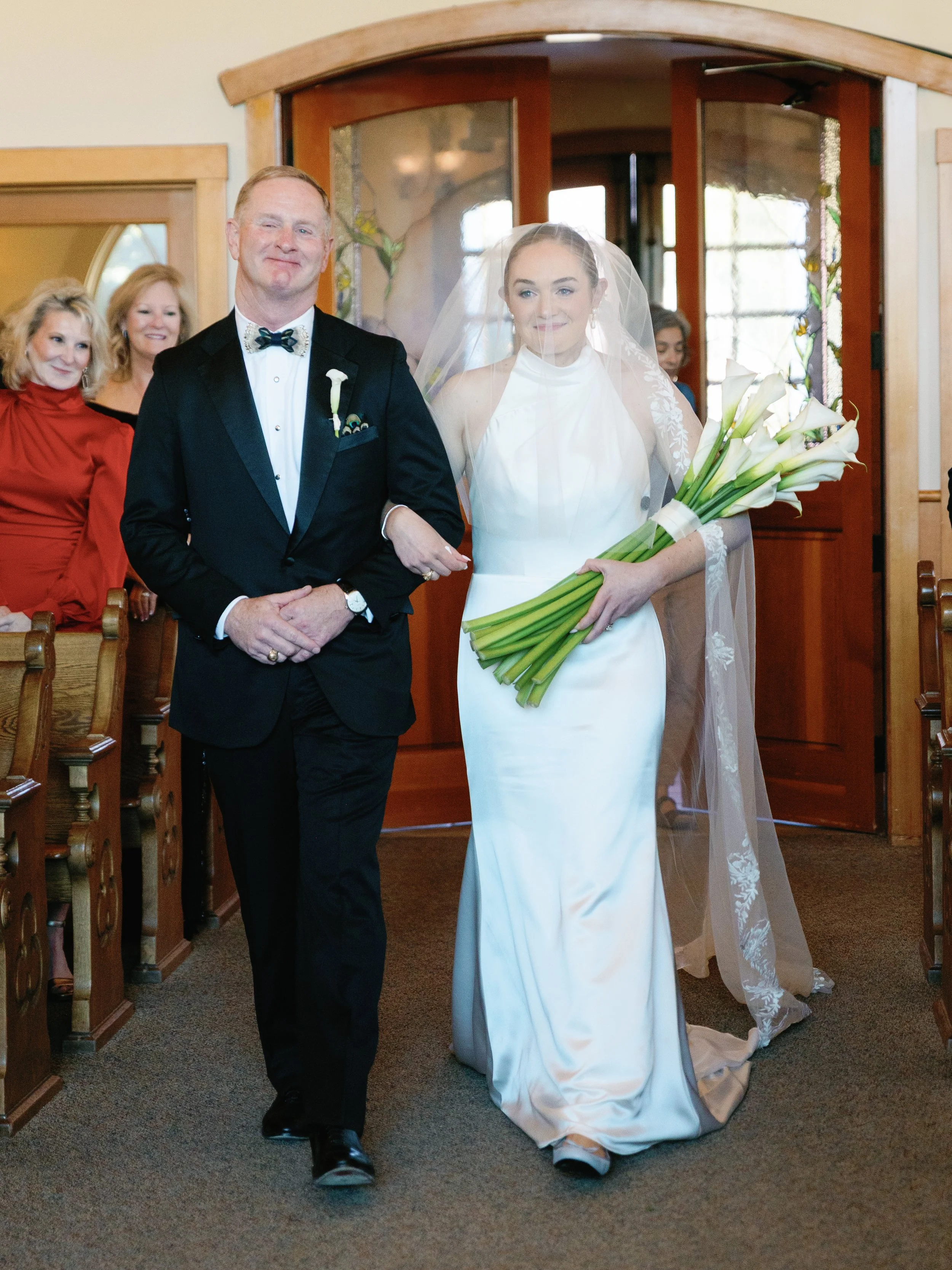 A bride in a white wedding dress holding a bouquet of calla lilies walking down the aisle with her father, surrounded by wedding guests.