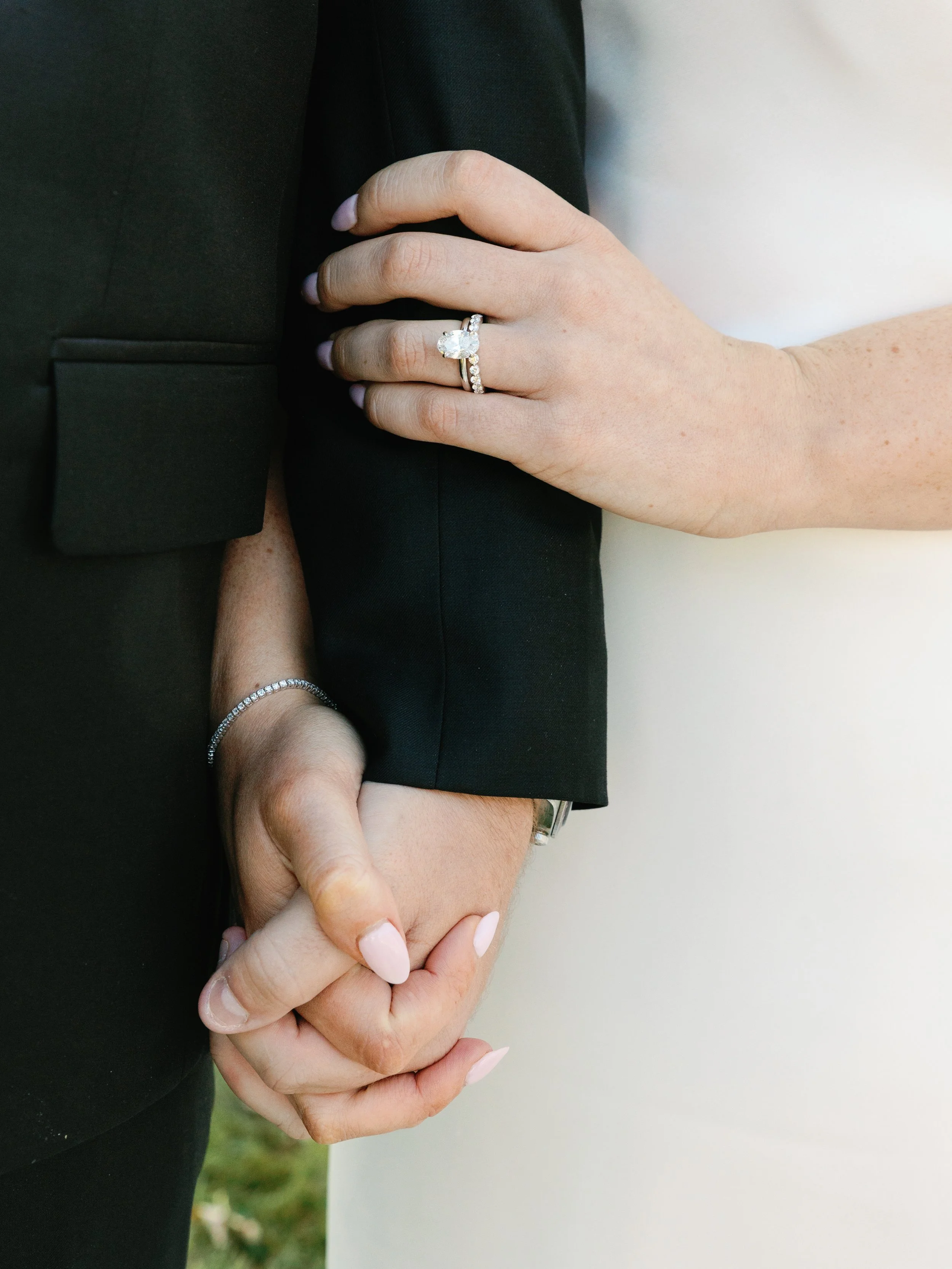 A bride and groom holding hands, with the bride wearing an engagement ring and wedding band, and the groom wearing a wedding band, against a blurred background.