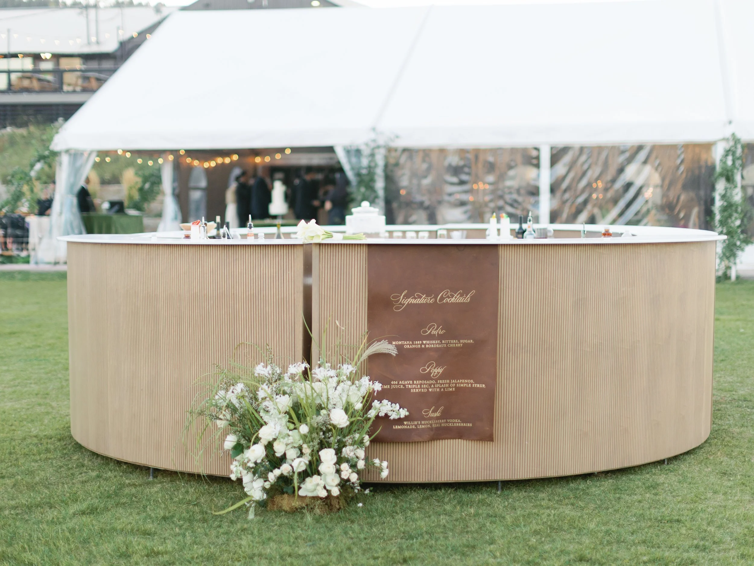 A bar counter with a flower arrangement in front, set outdoors at an event with a white tent in the background.