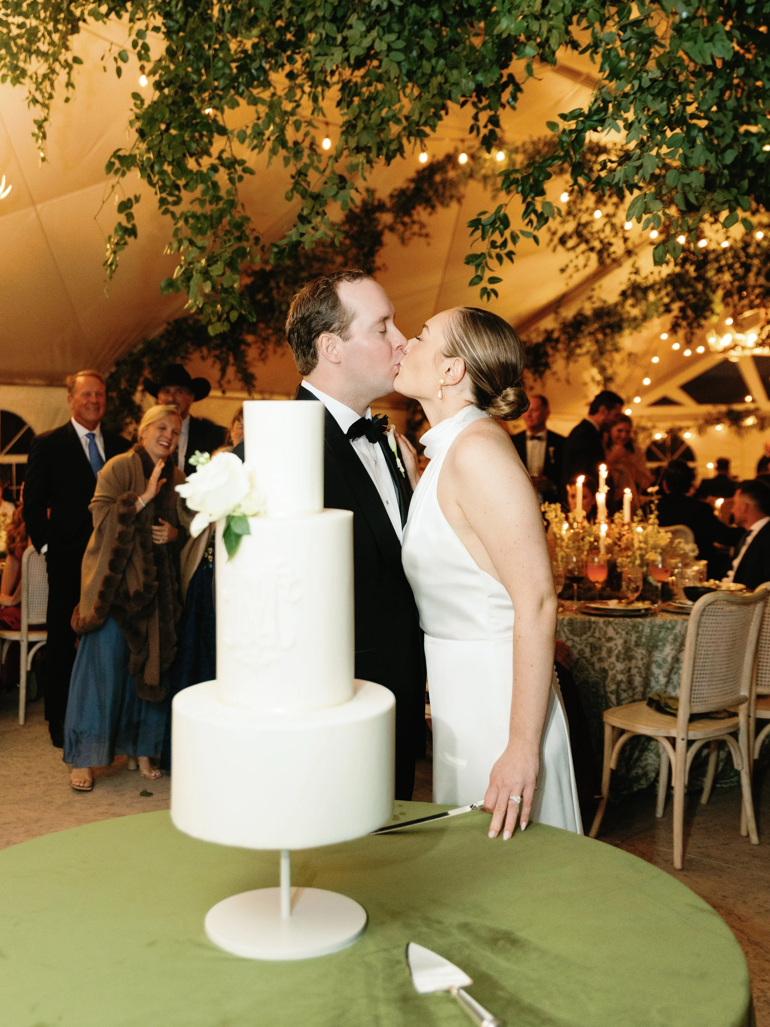 A bride and groom kiss at their wedding reception under a decorated canopy, with a wedding cake in the foreground and guests in the background.