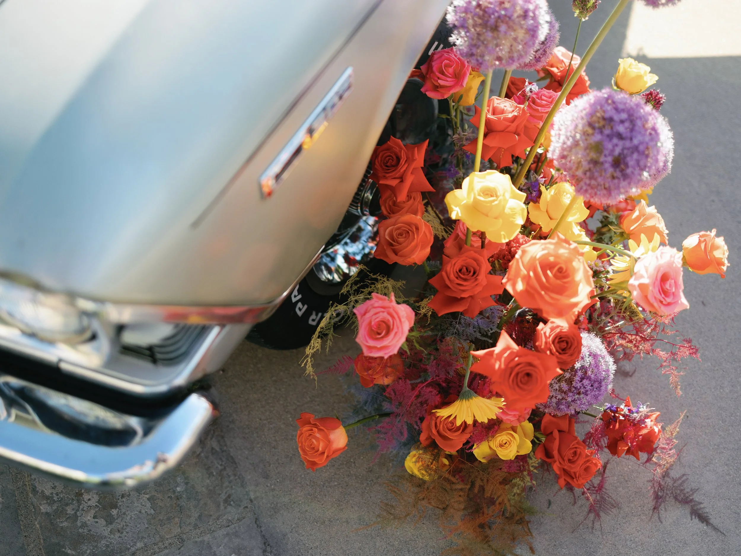 Close-up of a vintage car with a colorful bouquet of flowers, including roses and alliums, placed on the ground beside the car.