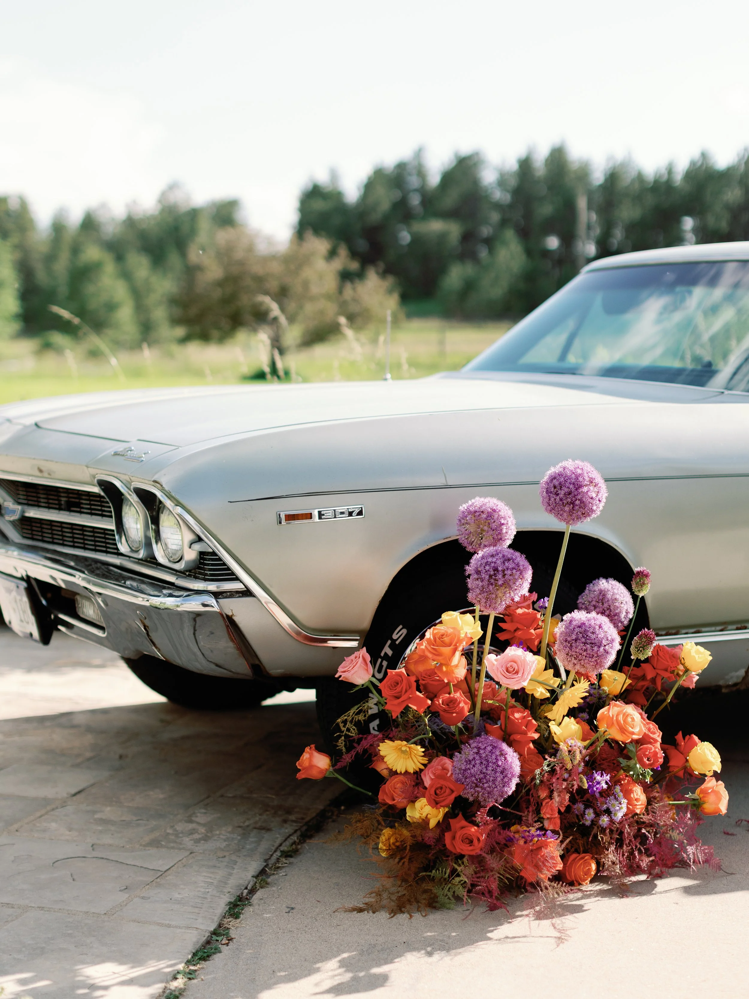 A vintage silver Chevrolet car with a floral arrangement of purple, orange, pink, and yellow flowers placed on the front wheel.