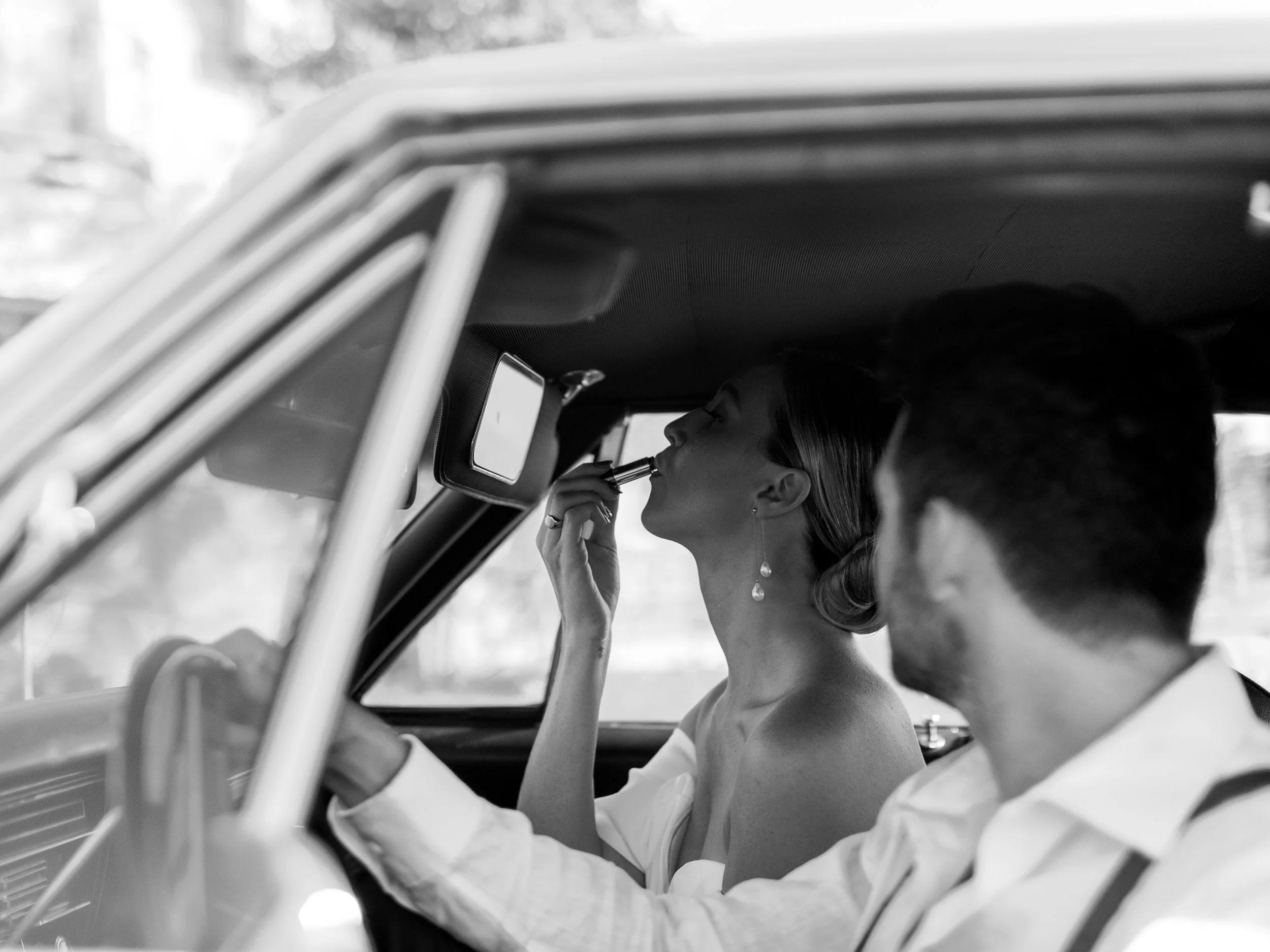 A woman sitting in a car, applying lipstick, with a man driving beside her.