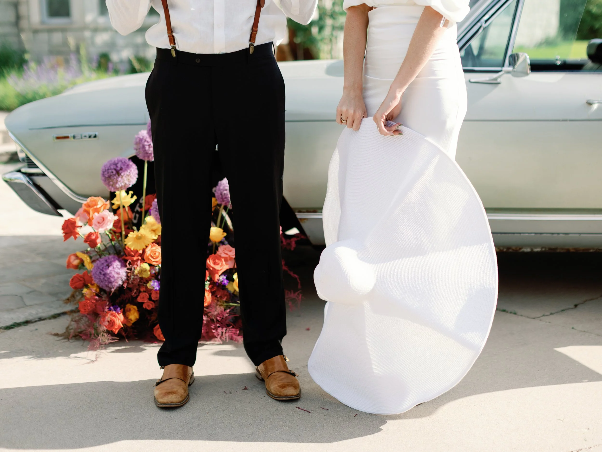 Partially visible couple dressed for a wedding with a vintage car and colorful flower arrangement in the background.