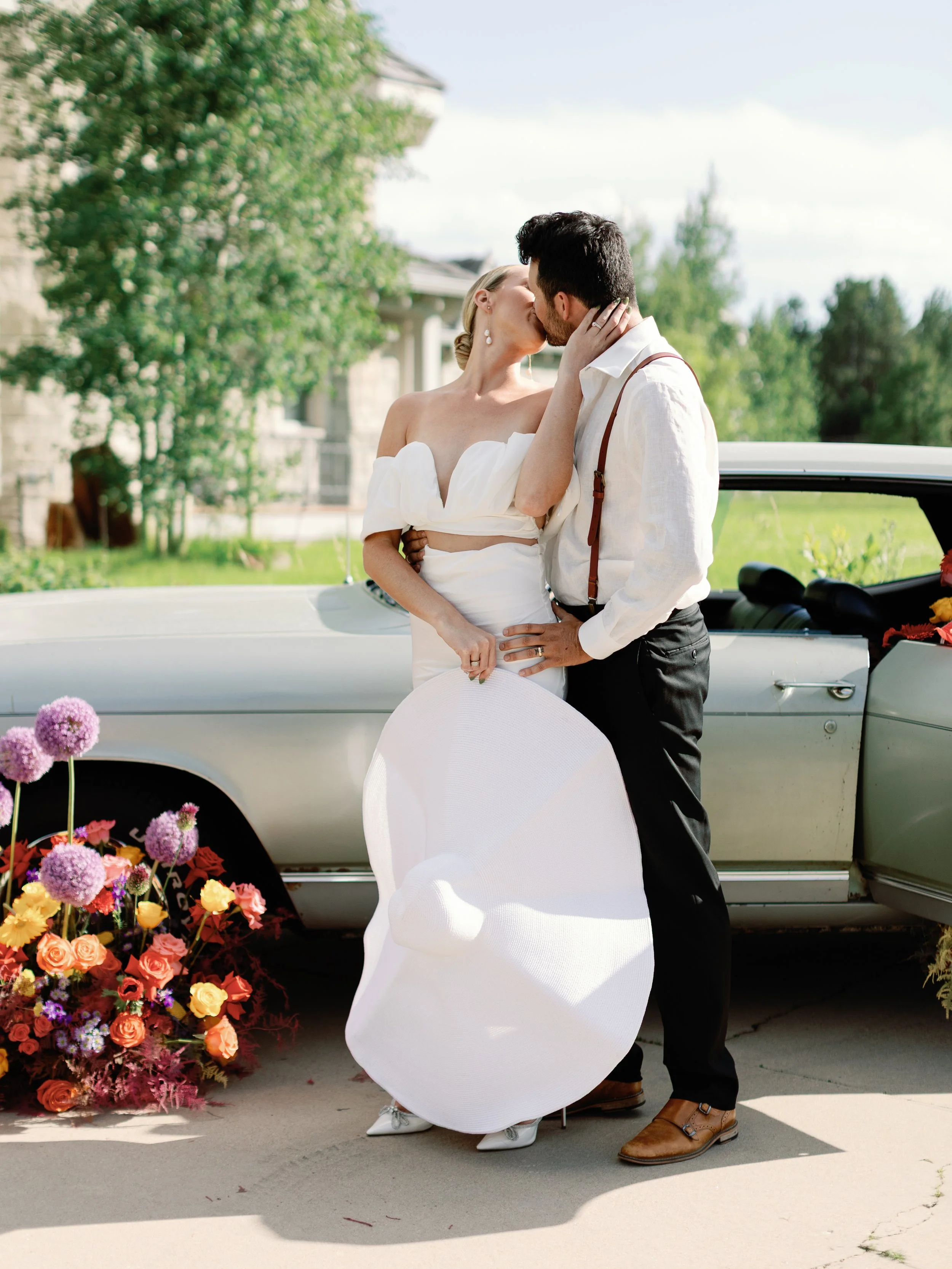 A couple dressed in wedding attire sharing a kiss outdoors near a vintage car decorated with flowers, with a bouquet of colorful flowers on the ground and a large white sunhat hanging from the woman's hand.