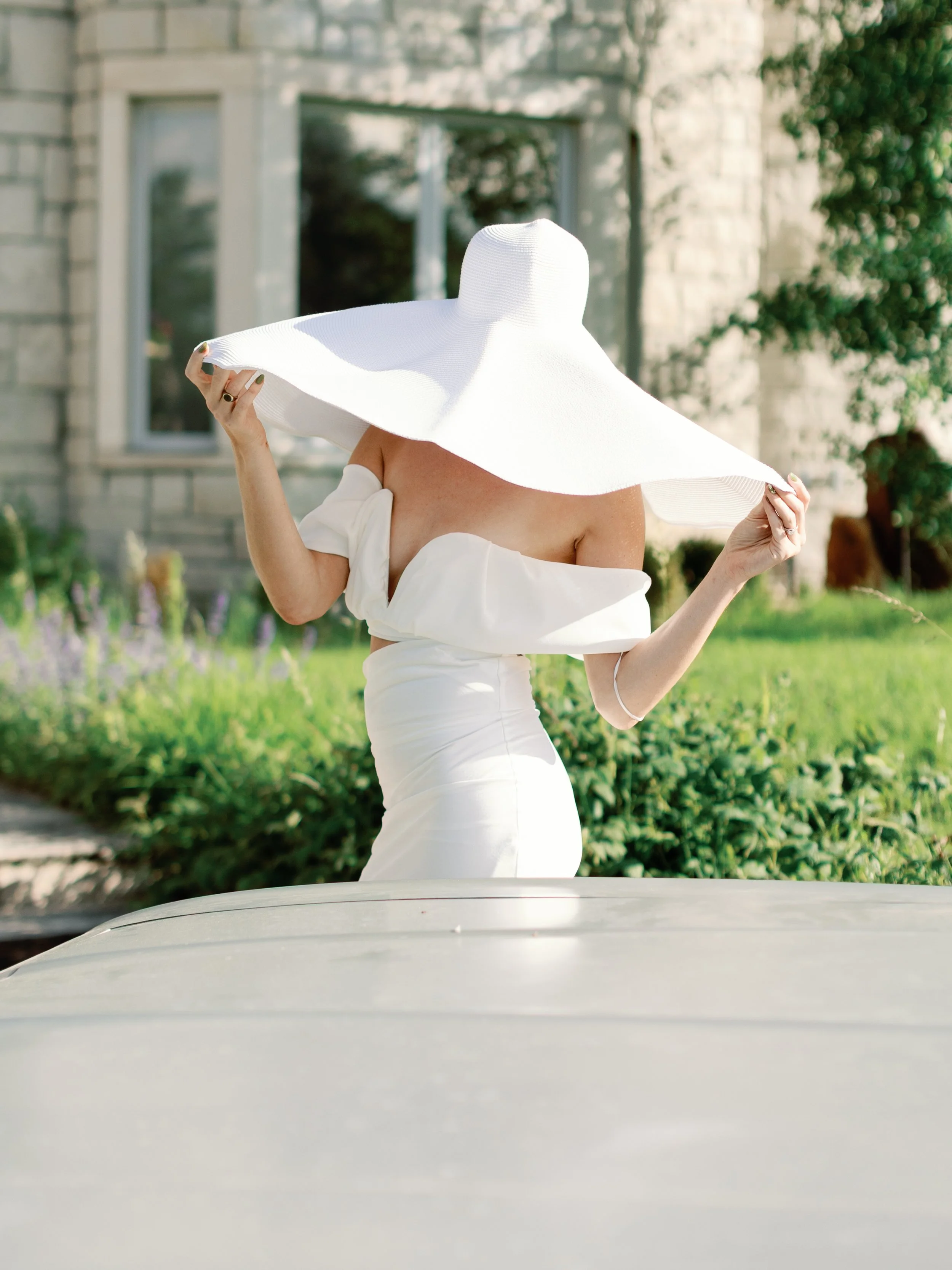 Woman in a white dress and large white sunhat standing outdoors in front of a stone building with greenery.