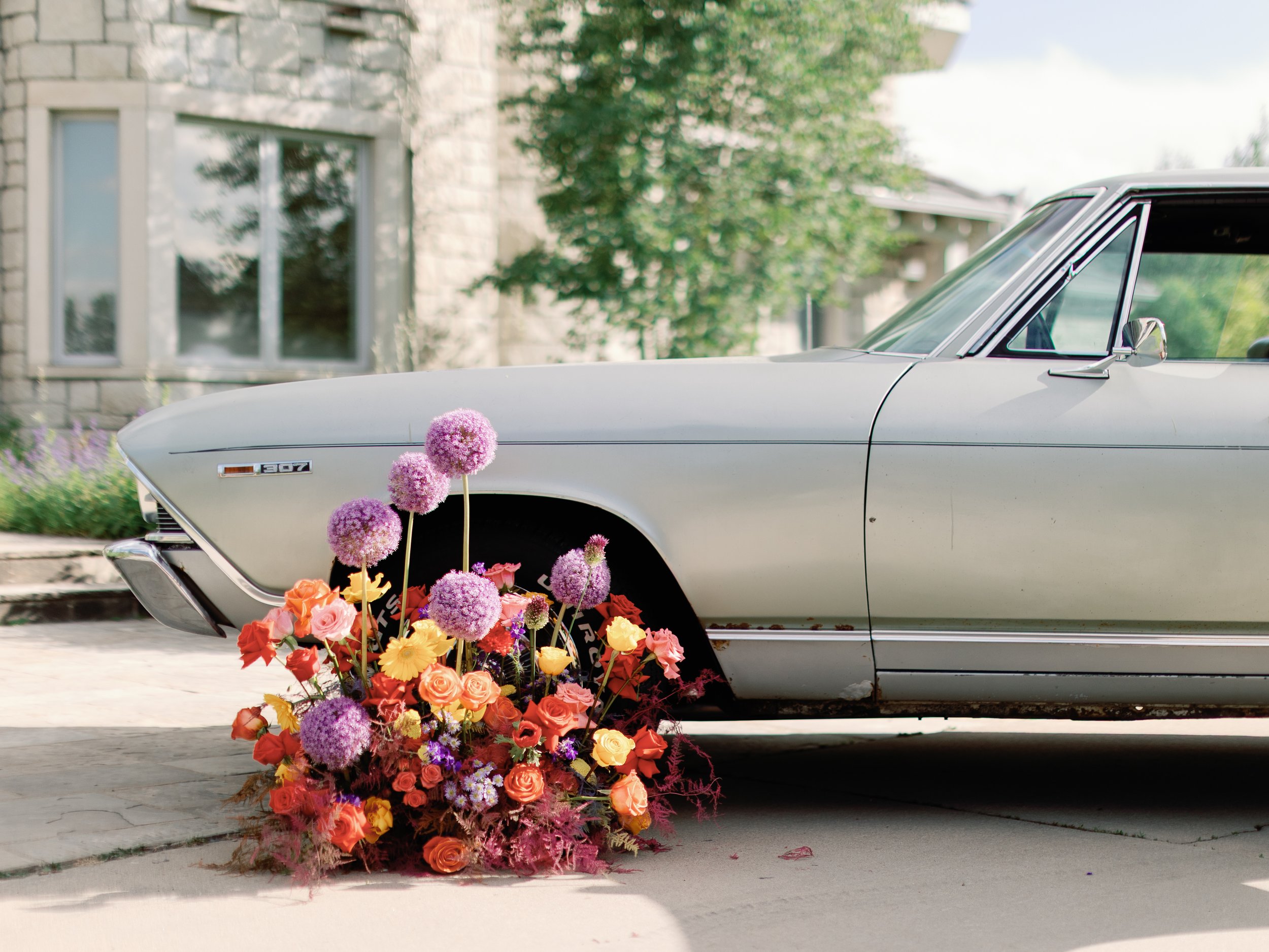 A vintage silver car with a flower arrangement at the front left wheel, including purple alliums and colorful roses, parked in front of a house with a stone facade and a garden with green plants and flowers.