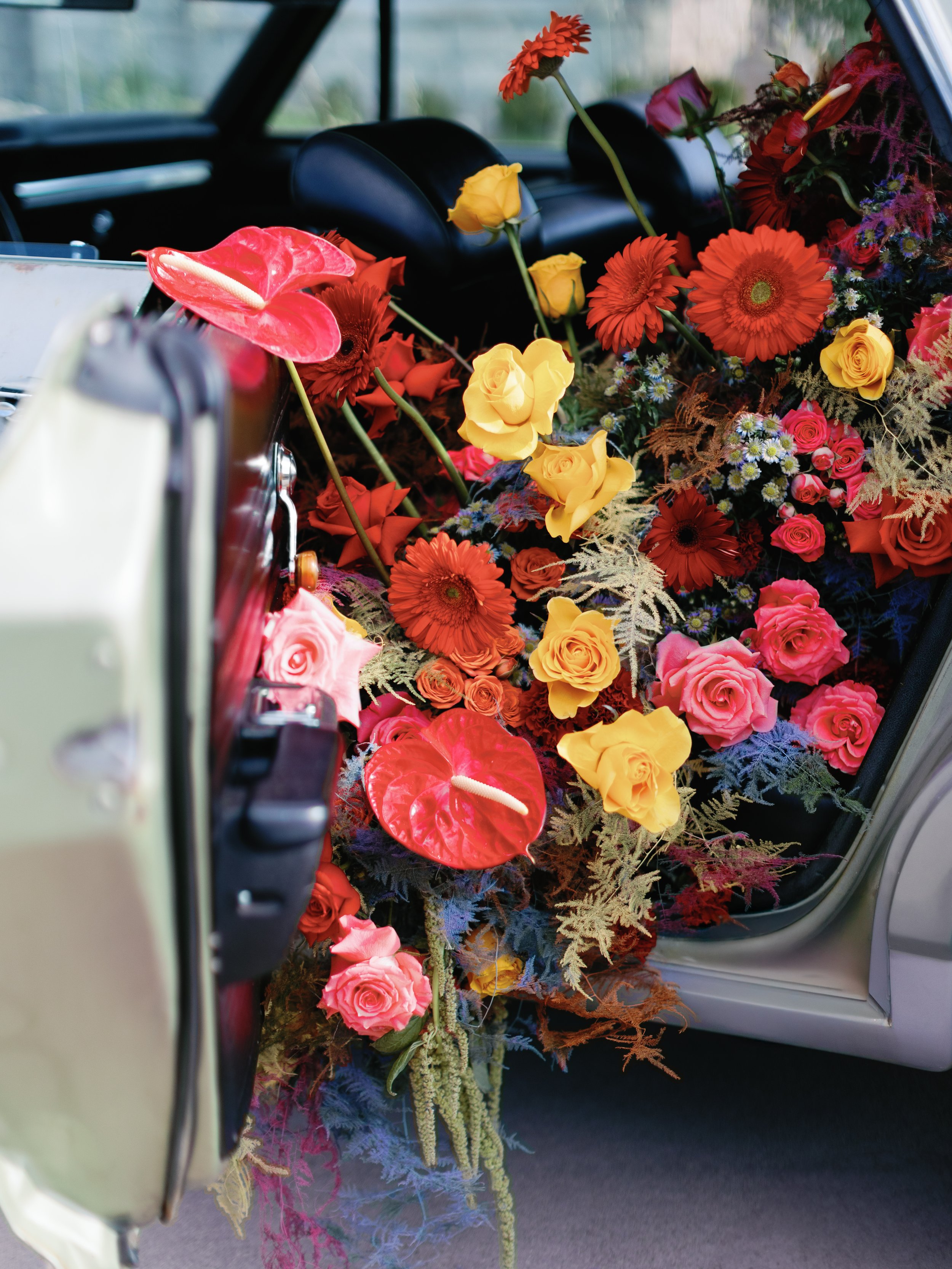 A car filled with colorful flowers including pink roses, yellow roses, red gerbera daisies, and red anthuriums, with some greenery and foliage, seen from the open back door.