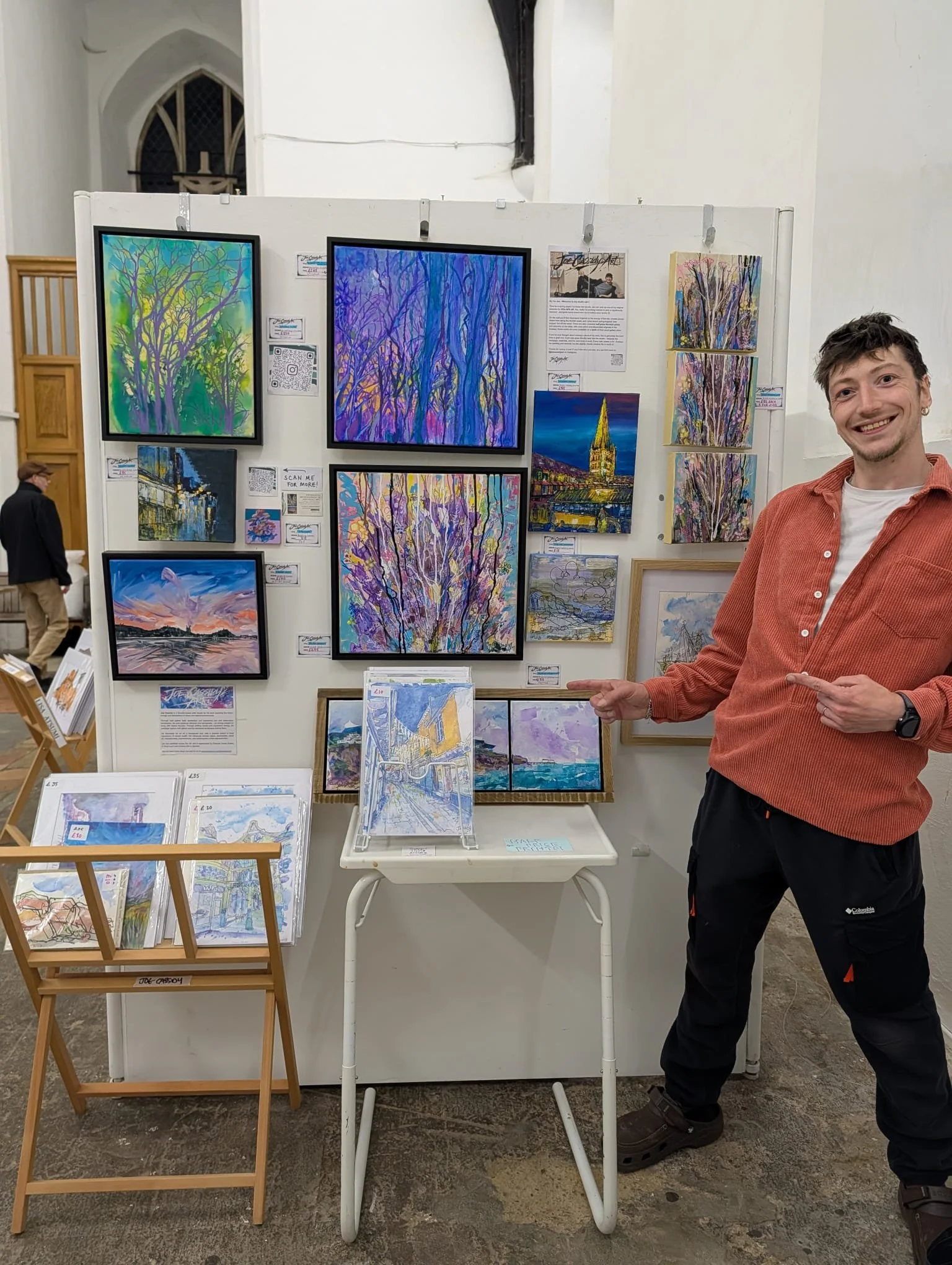 An art display booth with various colourful paintings of trees, a cathedral, and sunset scenes. A smiling man in an orange shirt points toward the artwork, standing in an indoor gallery or exhibition space.