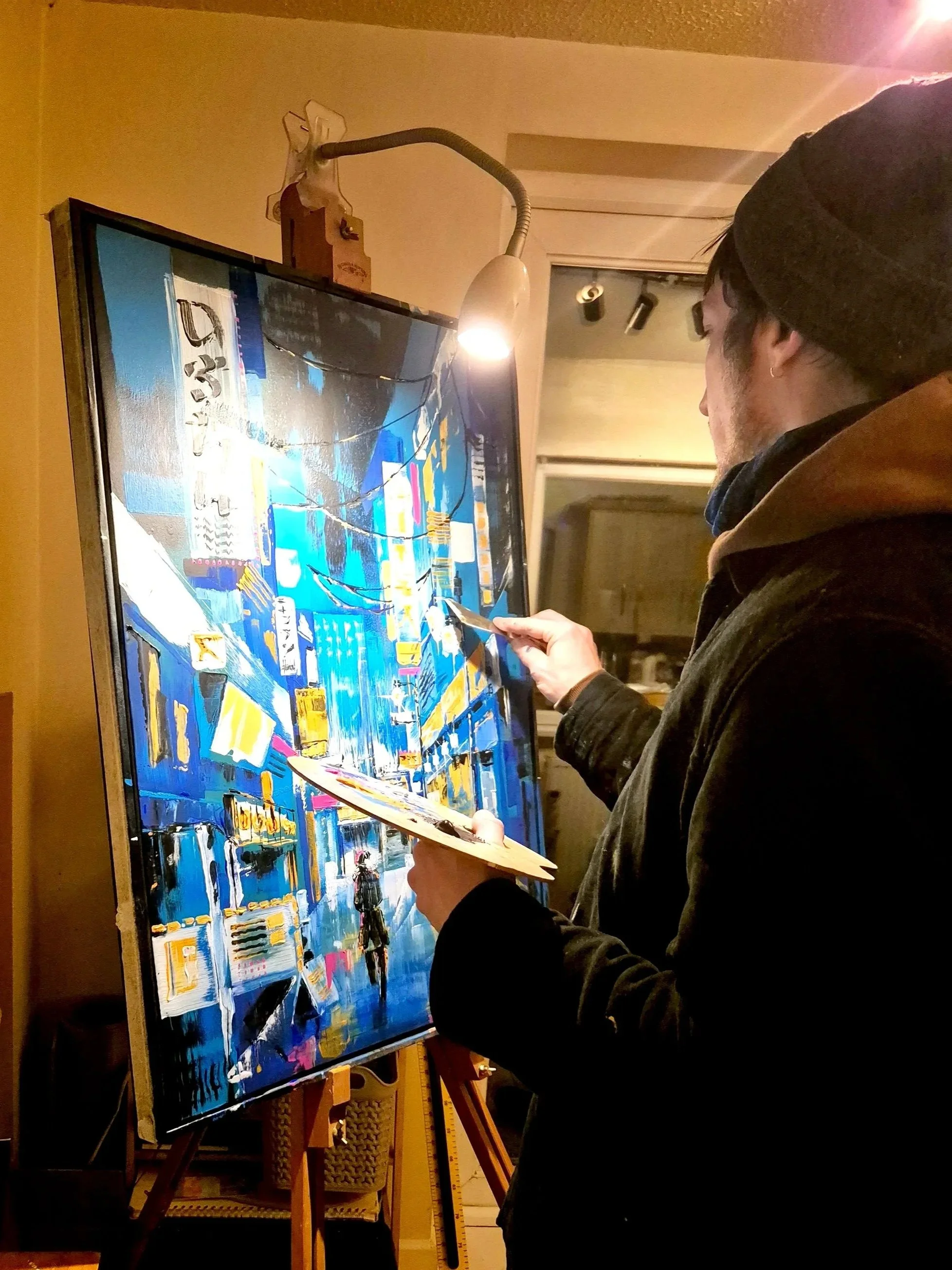 A person with dark hair, wearing a beanie and jacket, painting on a large canvas in an indoor space. The painting includes abstract and cityscape elements with vibrant blue, yellow, and white colours.