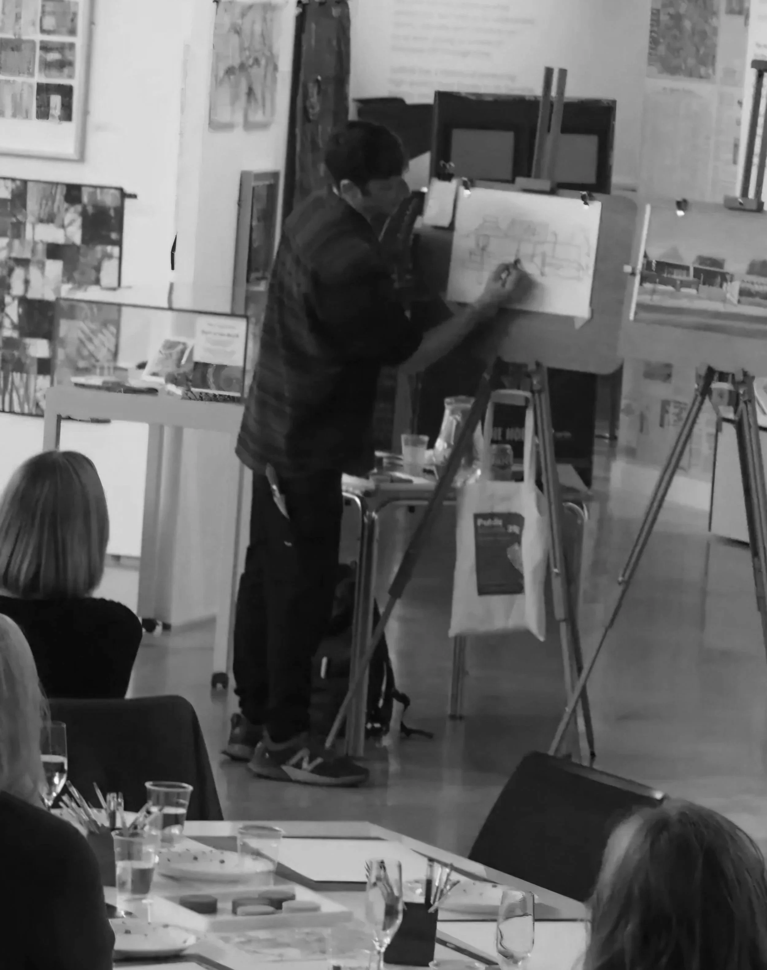 A person is drawing architectural sketches on a large sheet of paper in an art studio or classroom, surrounded by other artwork and art supplies.