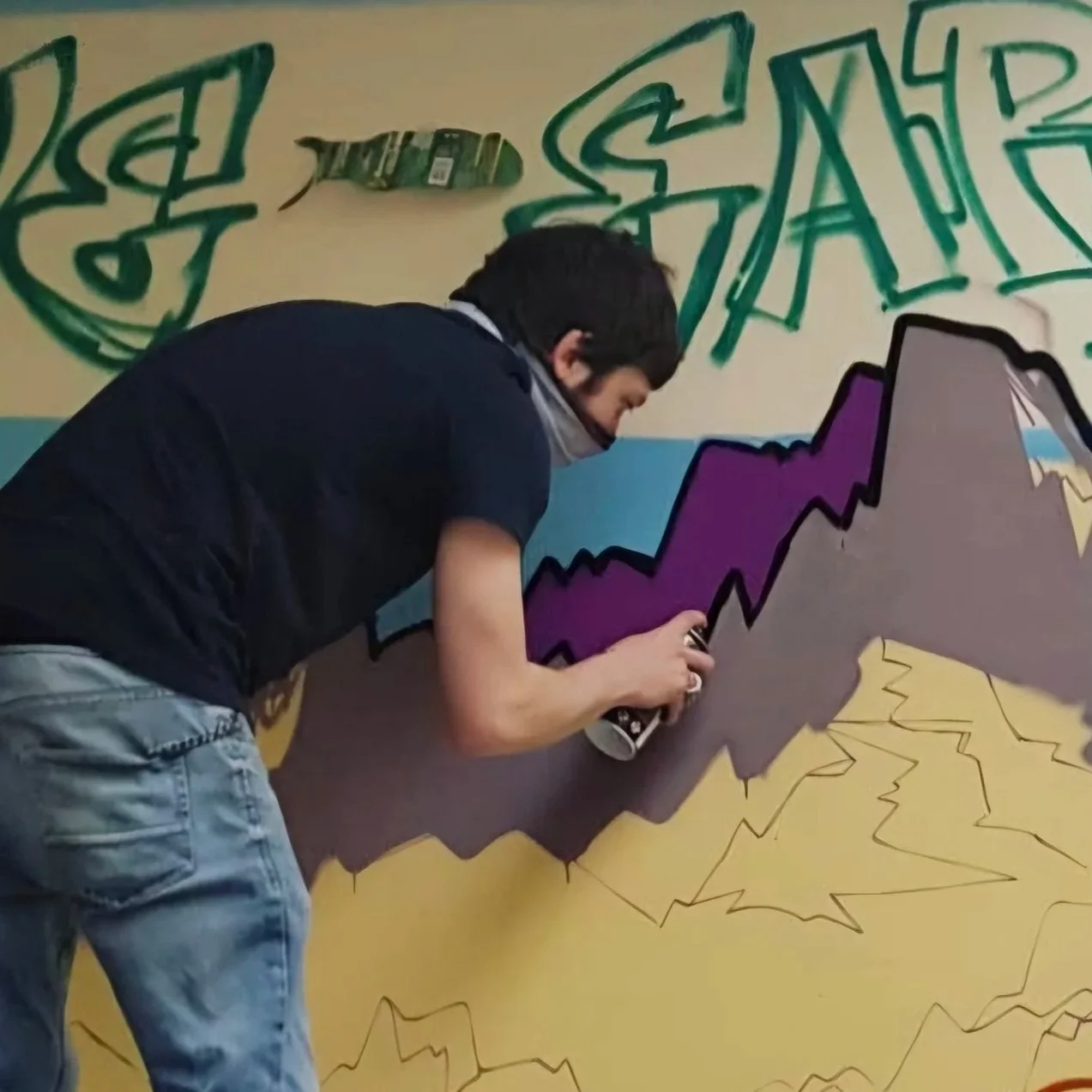 A person spray painting a purple mountain on a graffiti wall with a yellow background and green graffiti writing above.
