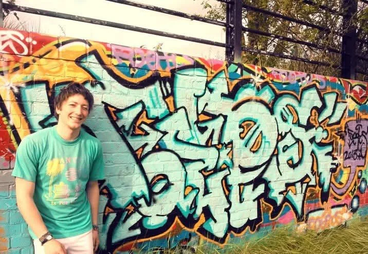 A young person with short hair smiling and wearing a teal T-shirt, standing in front of a colourful graffiti wall.
