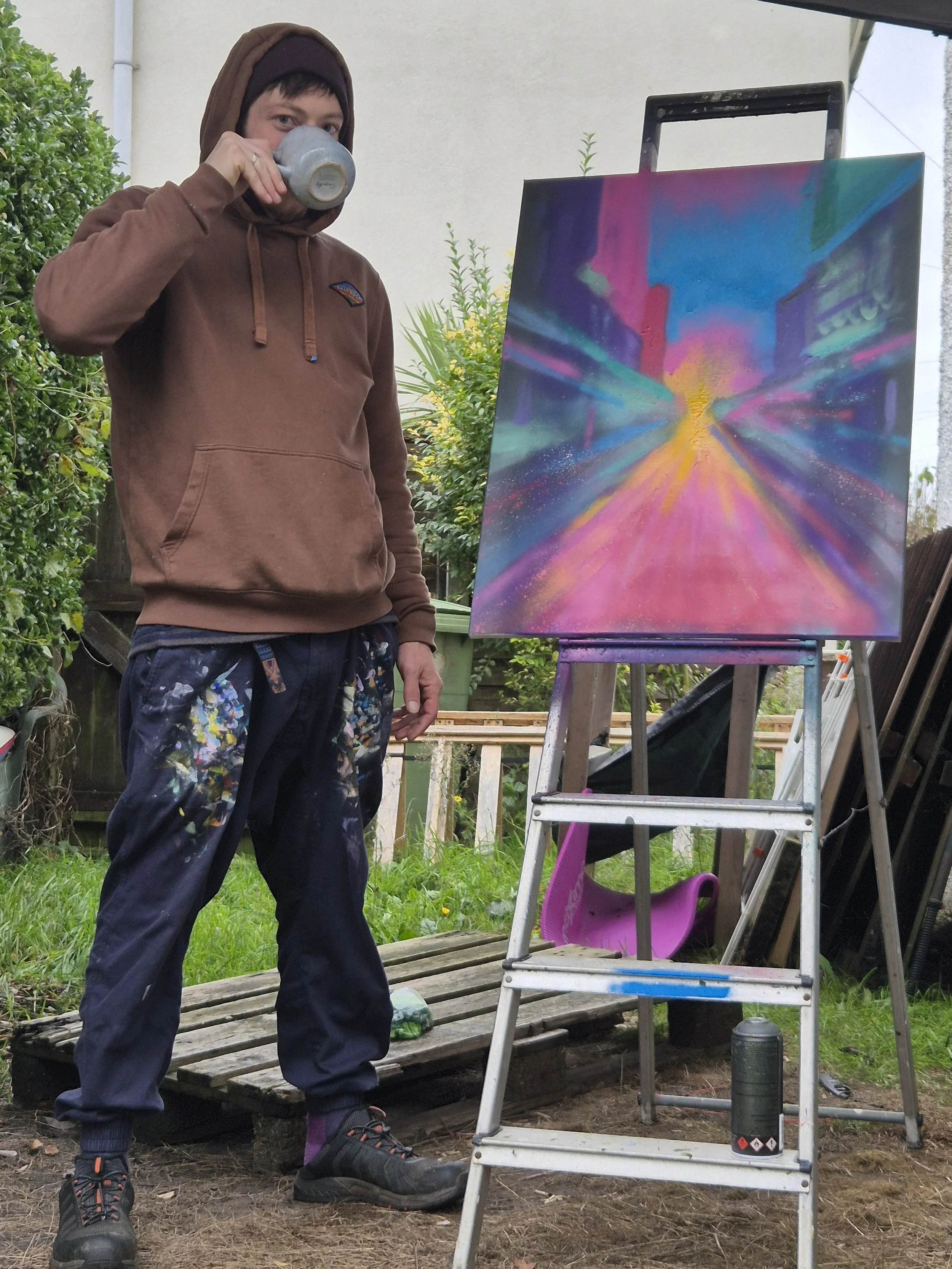 A young man in a brown hoodie and paint-splattered pants drinking from a gray mug outdoors, standing next to an easel with a colourful, abstract cityscape painting. The setting is a backyard with greenery and a wooden pallet on the ground.