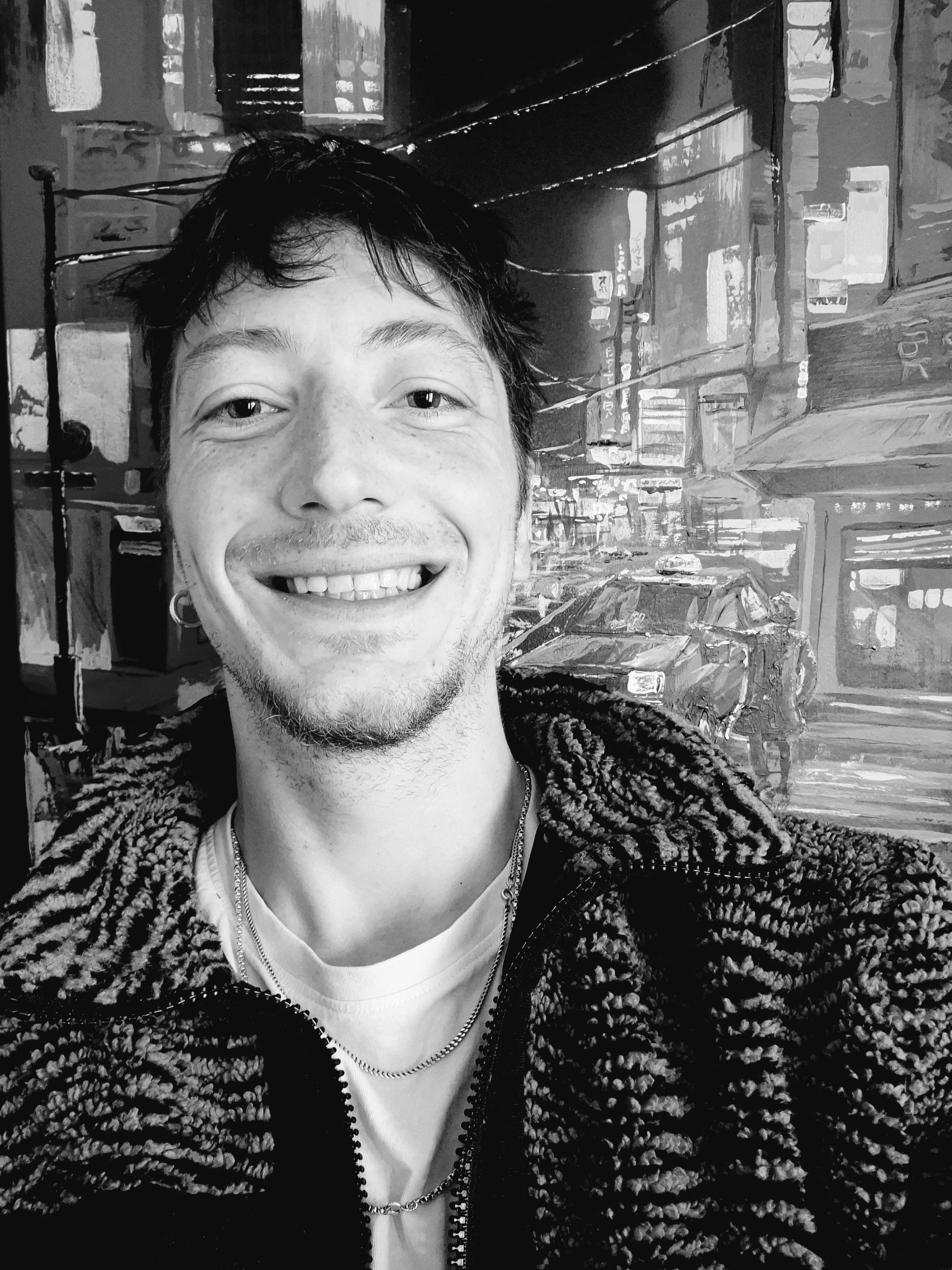 Black and white photo of a smiling man with short dark hair, wearing a textured jacket, a white T-shirt, a necklace, and earrings, in front of an urban cityscape with bright lights and tall buildings.