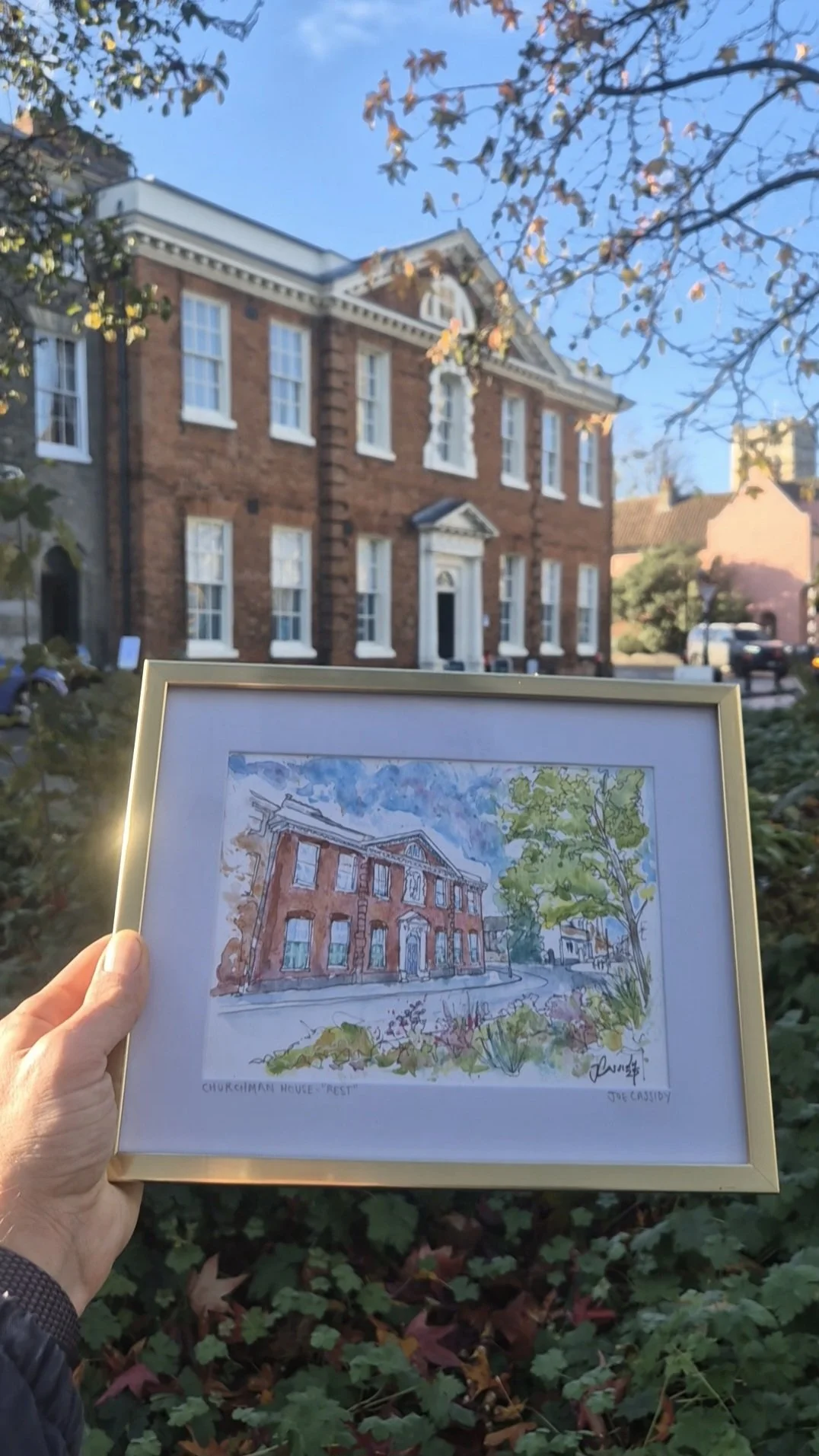 A person is holding a framed watercolor painting of the historic Churchman House, titled "Rest," in front of the actual brick house building that it depicts, with a bright blue sky and autumn leaves in the background.