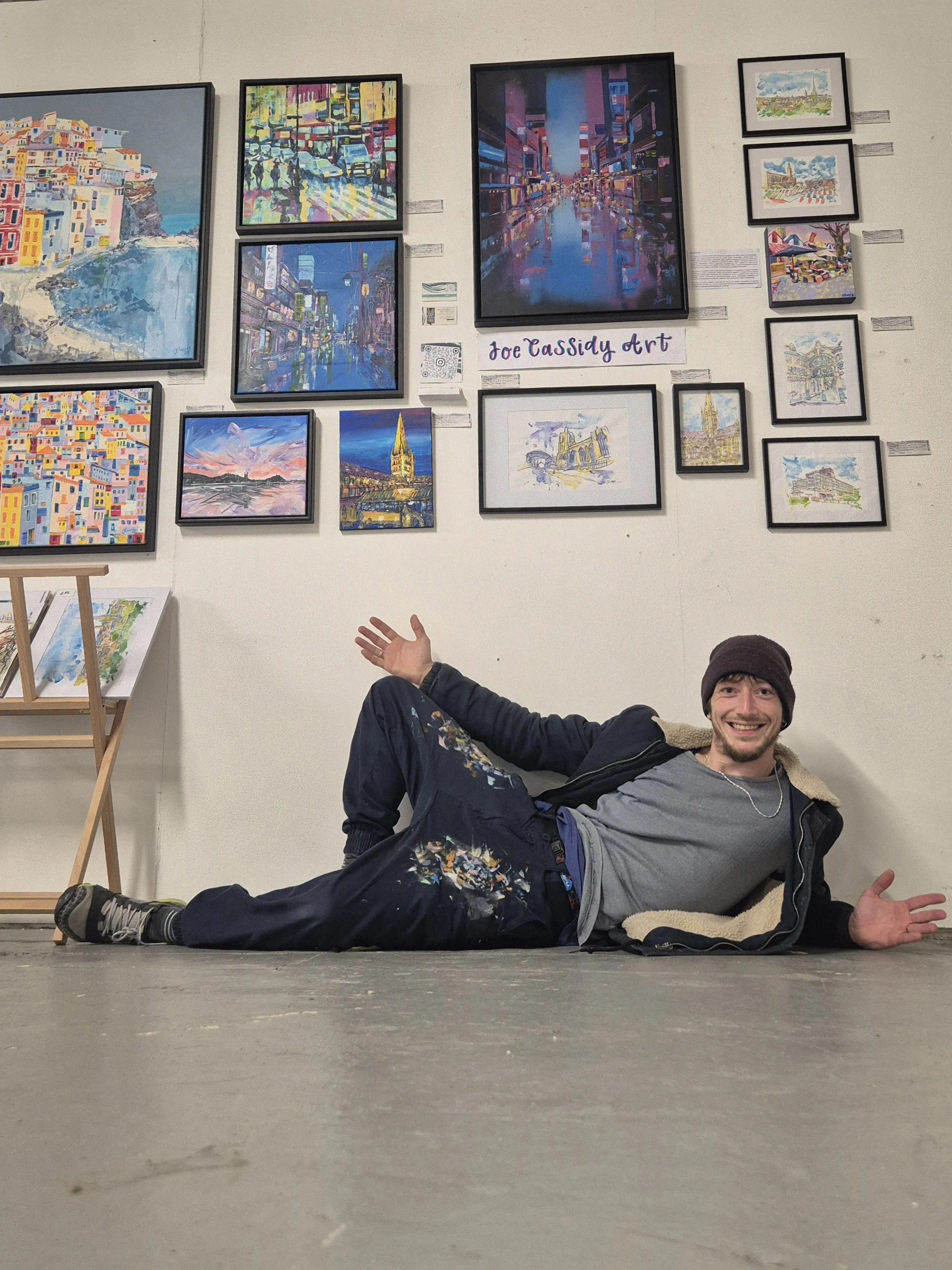 An artist lying on the ground in front of his artwork displayed on a gallery wall, smiling and making a thumbs-up gesture.