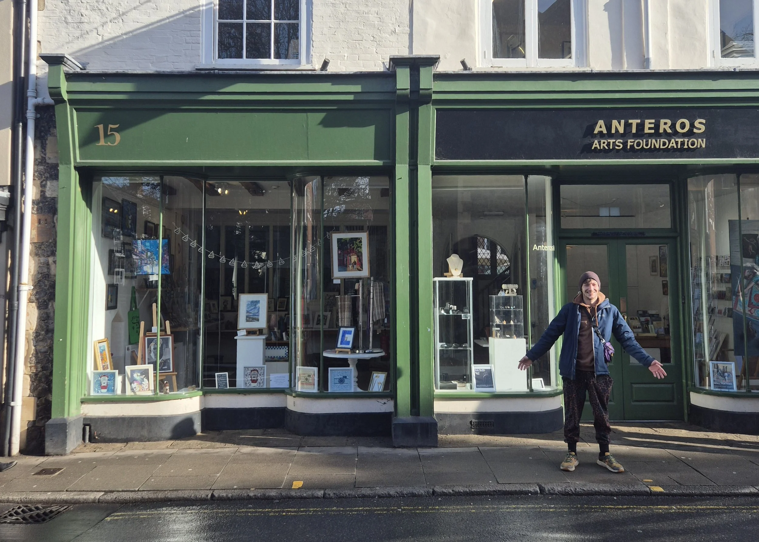 Storefront with large display windows showcasing art pieces, with green framing, street number 15, and a sign for Anteros Arts Foundation. A resident artist at the shop, Joe Cassidy, stands outside.