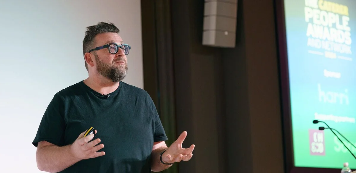 Man with glasses and beard giving a presentation at an awards event, standing next to a screen with the event's title and logo.