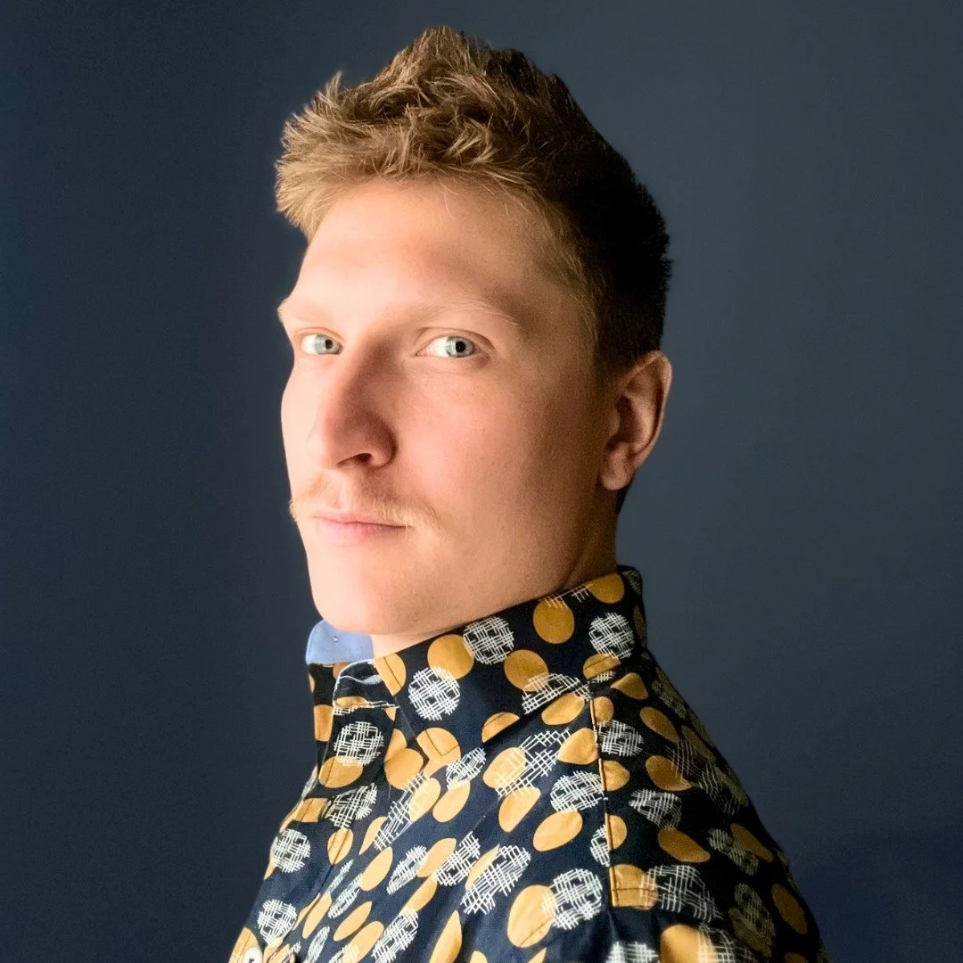 A young man with blonde hair and blue eyes, wearing a patterned shirt with yellow and black circles, looking at the camera against a dark background.