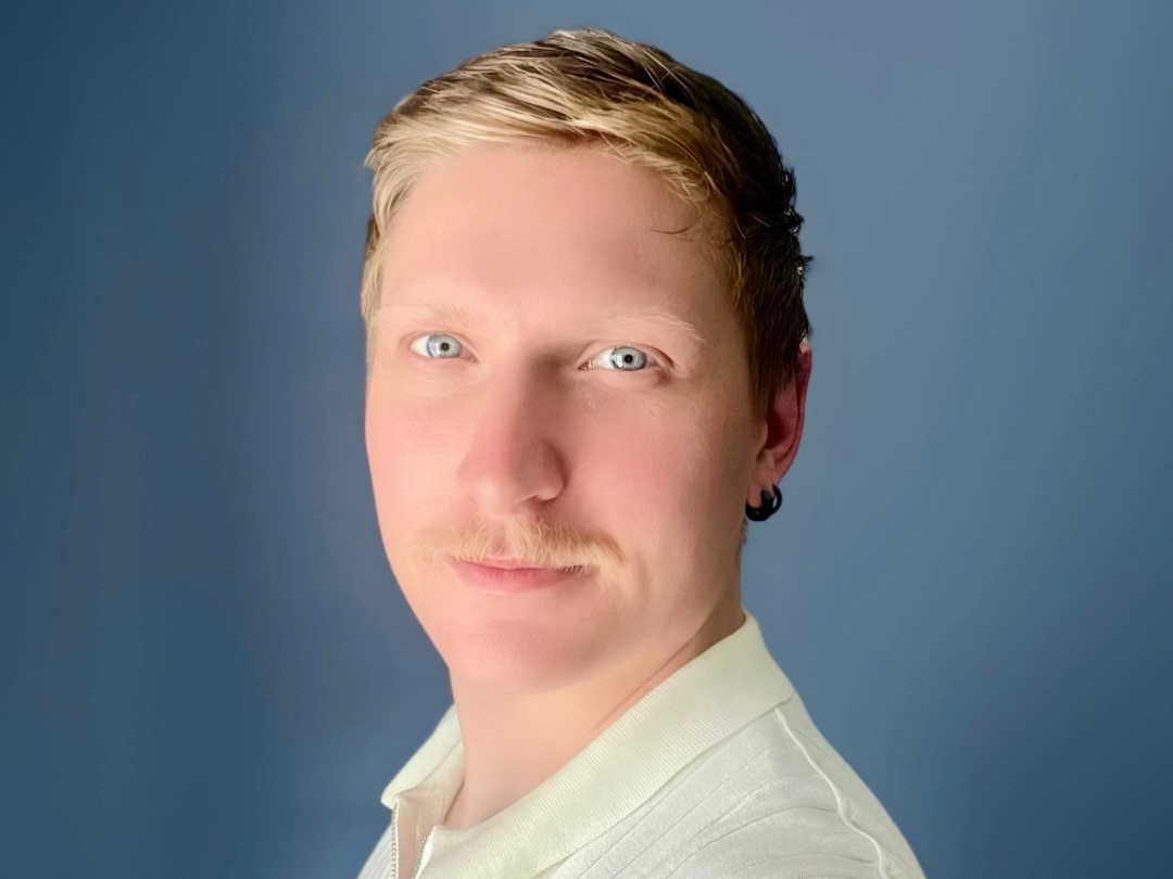 A young man with blonde hair, blue eyes, and a light mustache, wearing a white shirt and a small black earring, looking at the camera against a plain blue background.