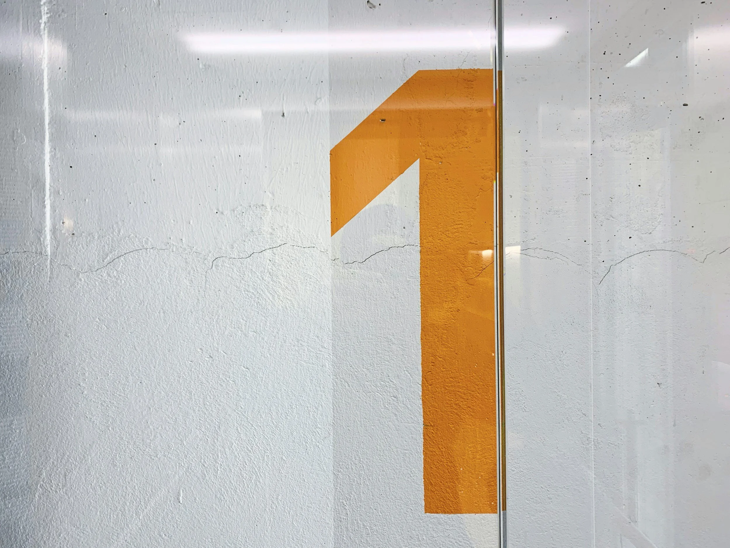 Large, orange arrow painted on a white wall, pointing upward. Part of the wall appears to be a mirror reflecting the surroundings.