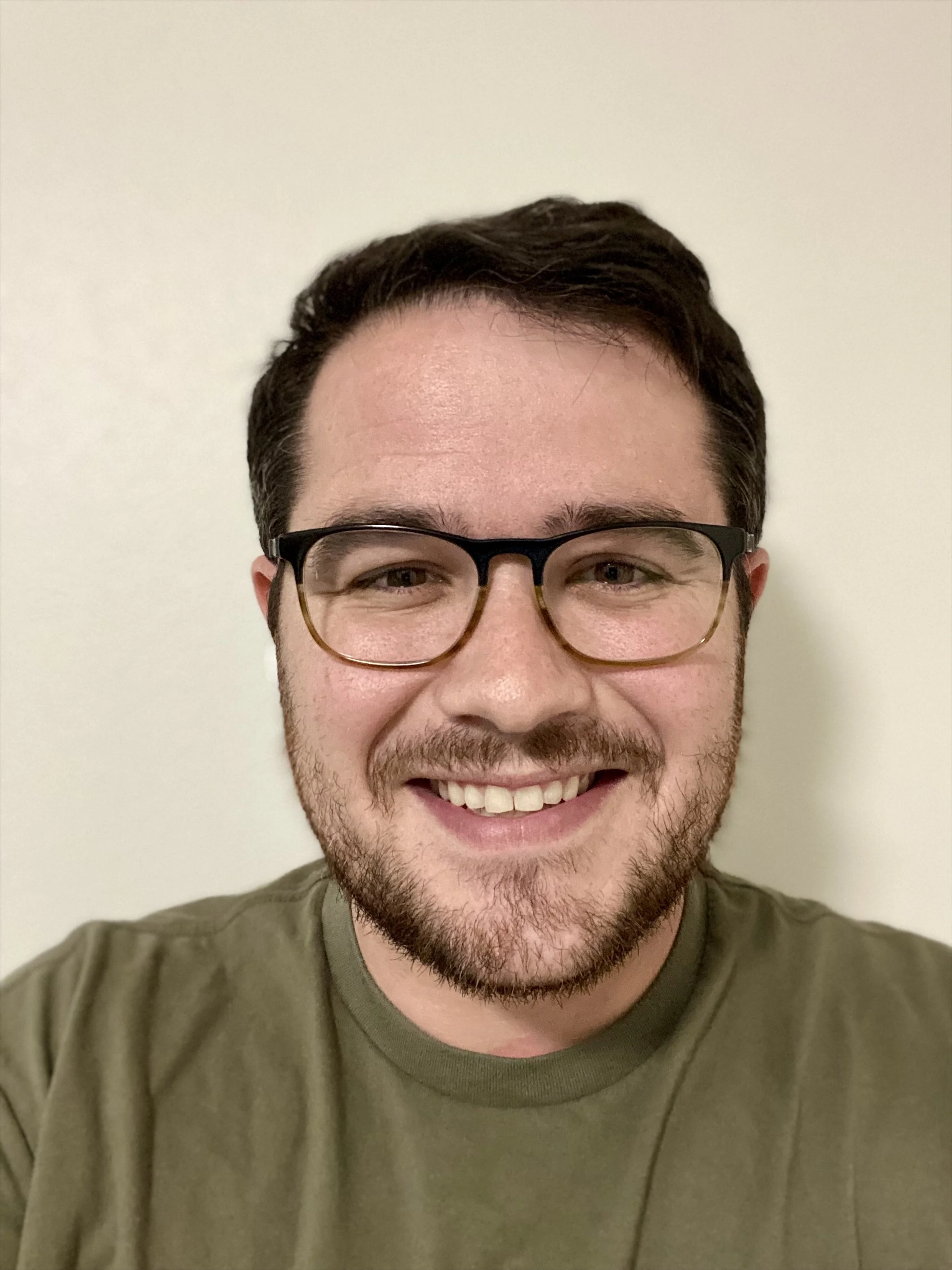 A young man with short dark hair, glasses, and a beard, smiling at the camera, wearing a green t-shirt, standing against a plain white wall.
