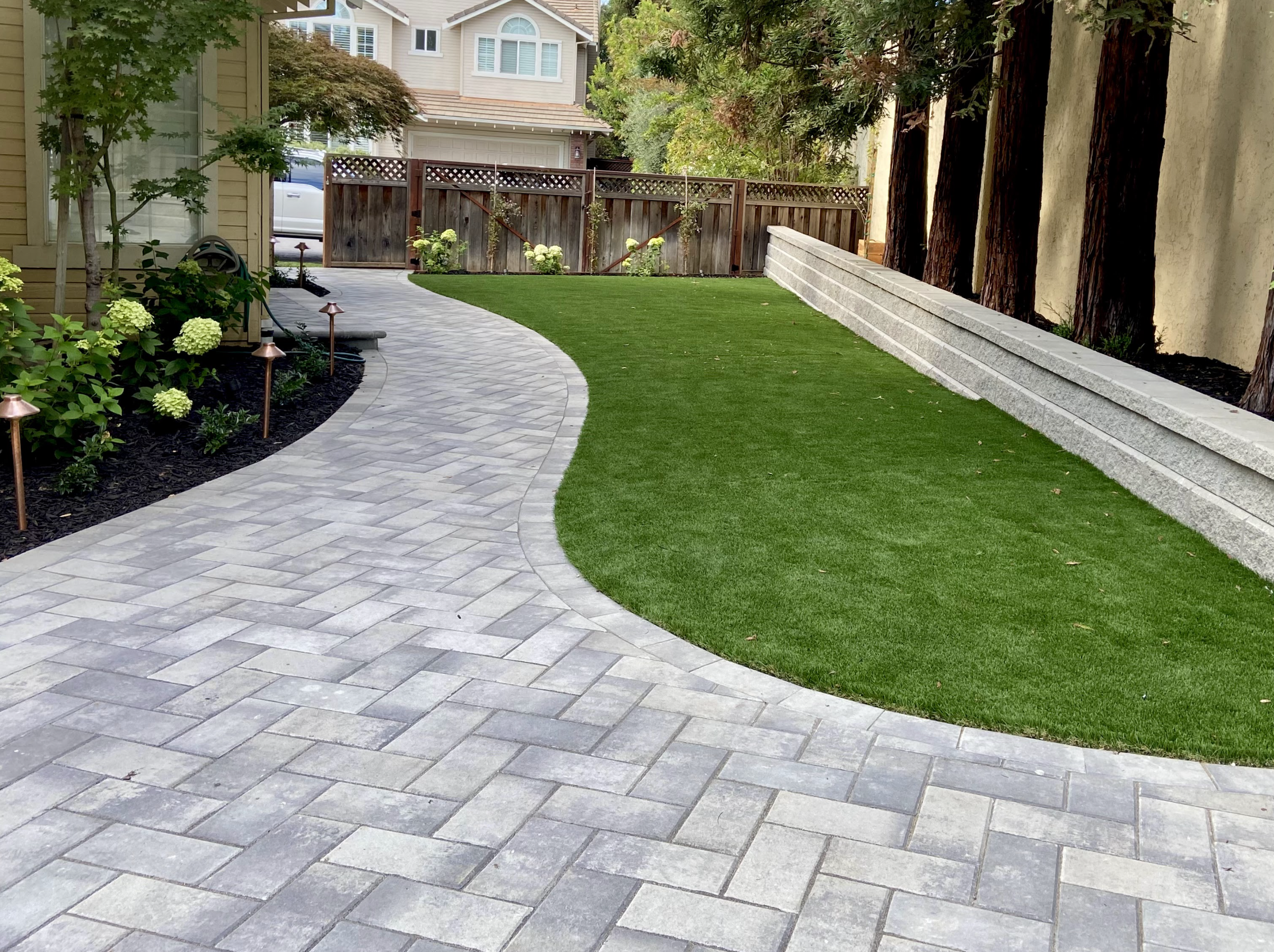 Decorative backyard with a curved stone pathway, green lawn, flower bed with white hydrangeas, and a wooden fence in the background.