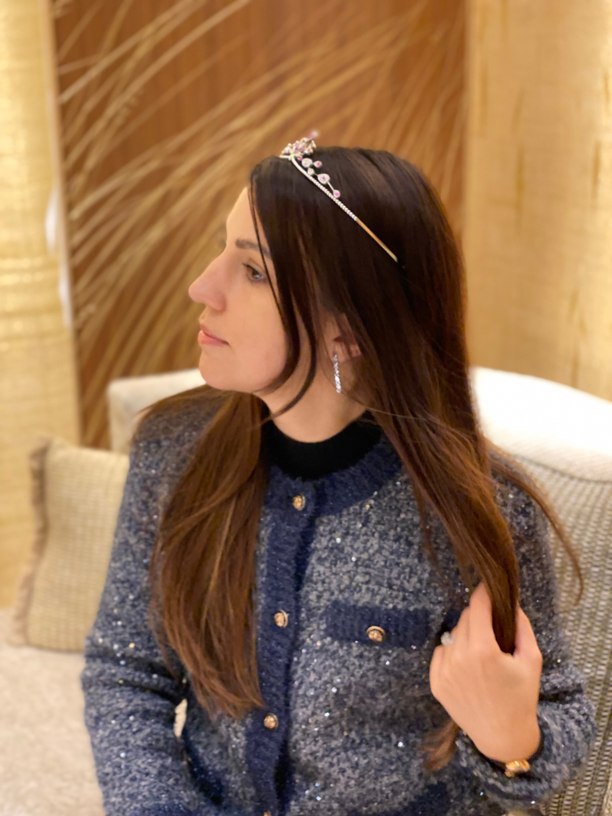 A woman with long brown hair, wearing a tiara, earrings, and a glittery gray jacket, sitting indoors with a decorative wooden background.