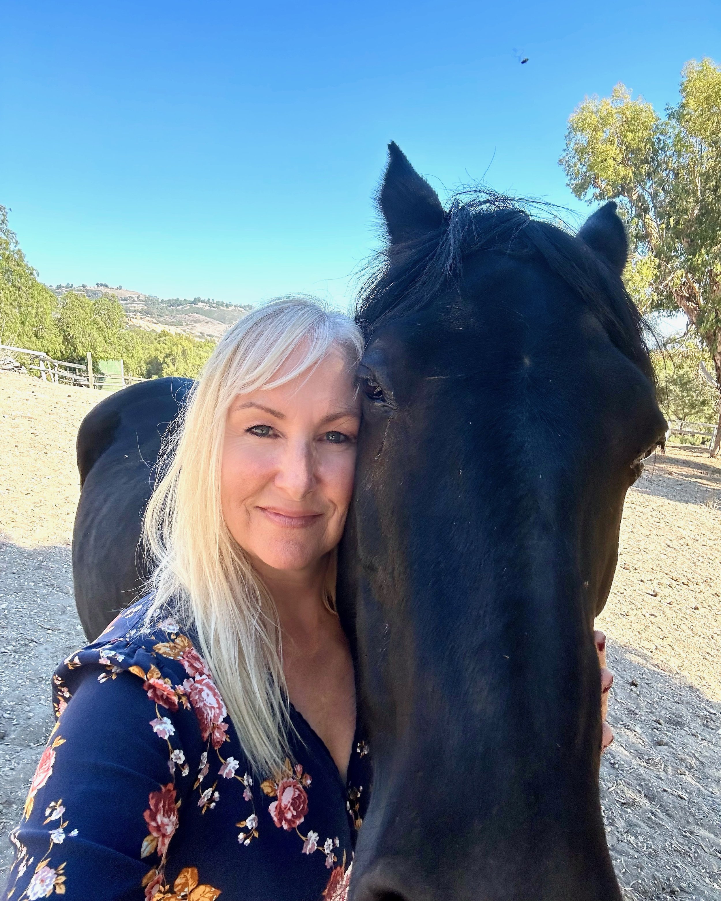 A woman with blonde hair and a black top with floral pattern hugs a black horse. The horse has its ears pointed forward and is close to the woman’s face. The background features a clear blue sky, trees, and a distant hillside.
