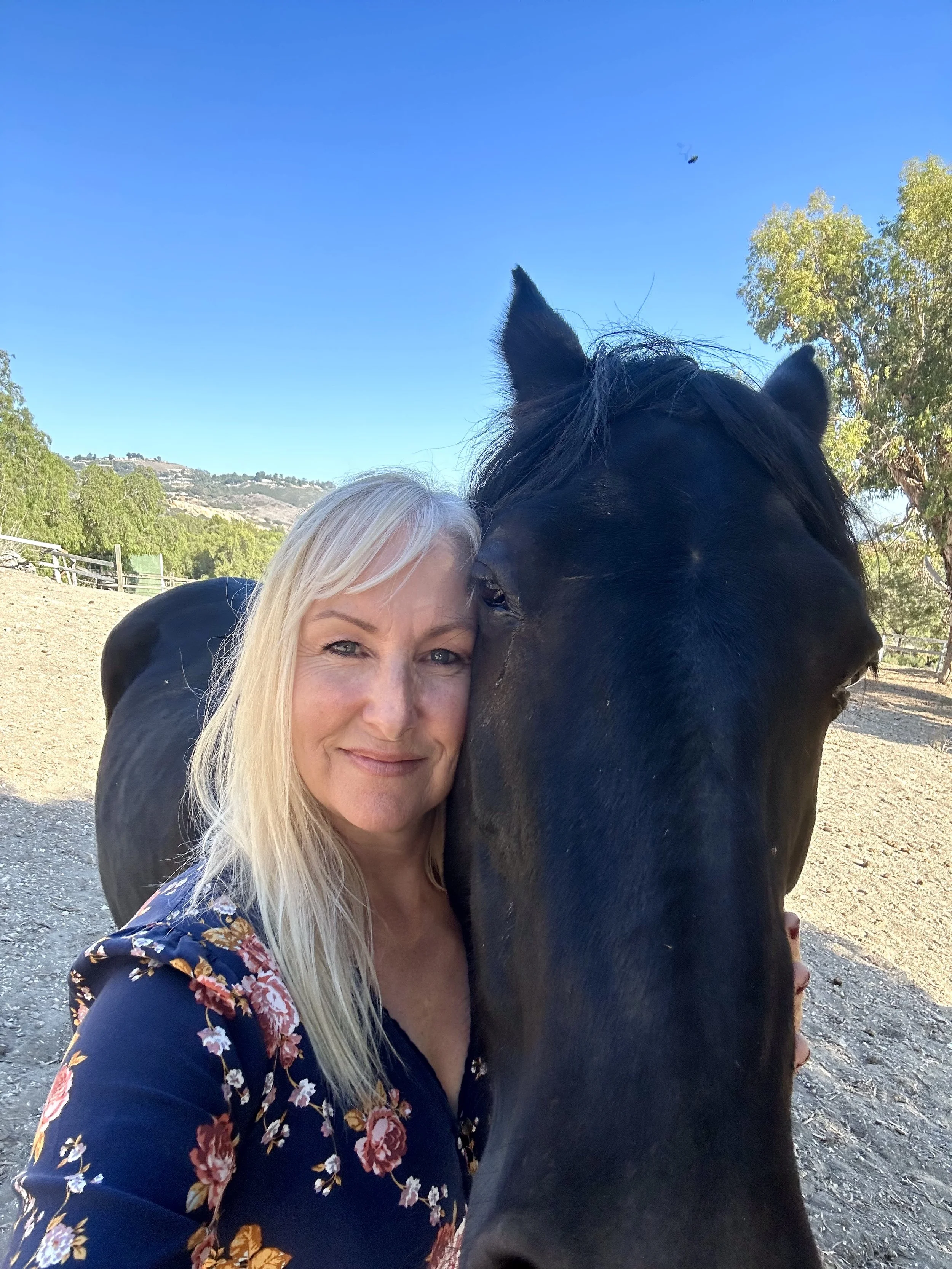 A woman with long blonde hair leaning her head against a black horse, both appearing happy outside on a sunny day with trees and hills in the background.