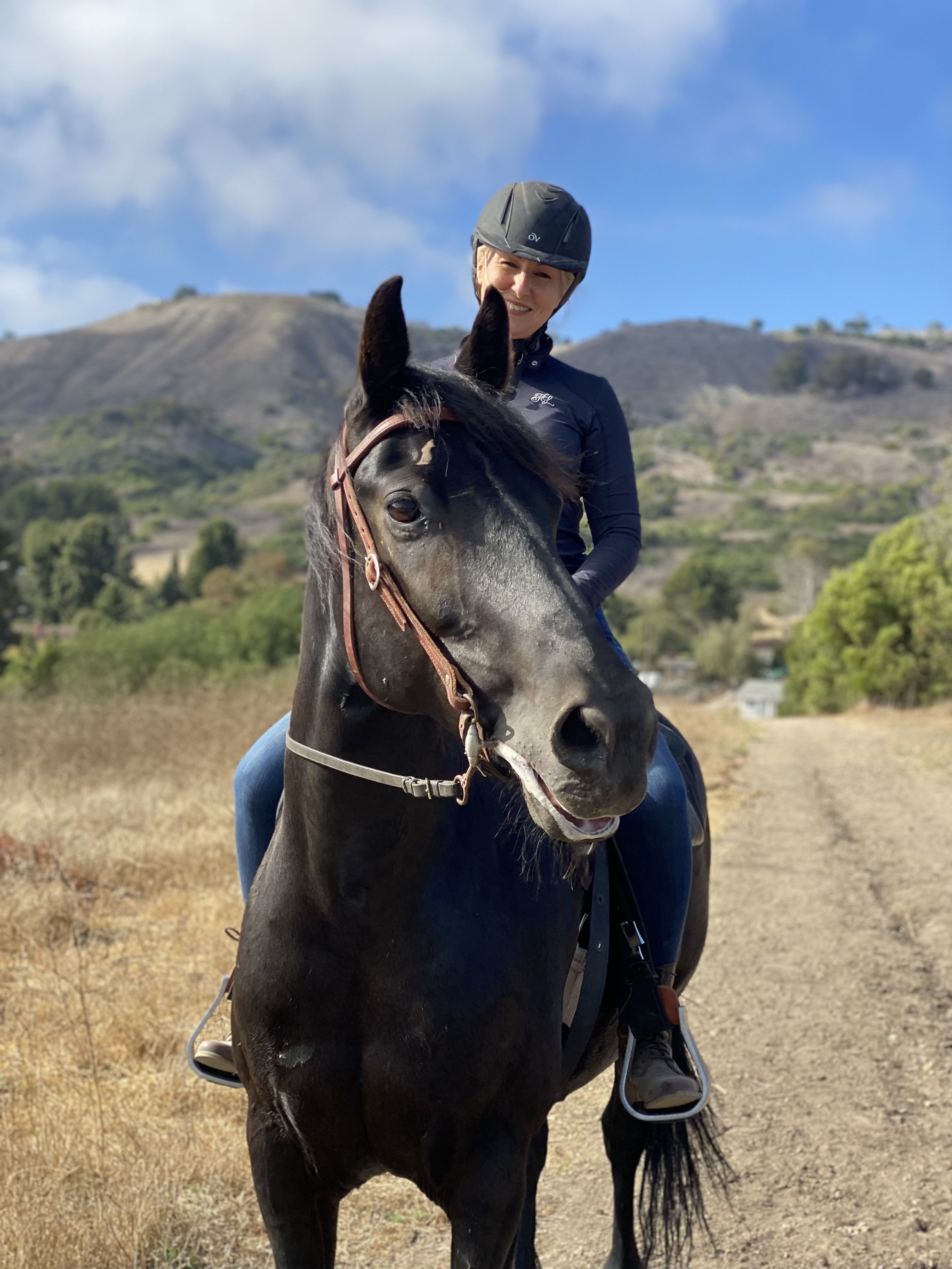 A woman wearing a helmet riding a black horse in an open field with hills and trees in the background.