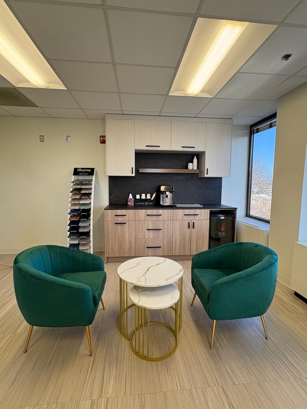 Small office kitchenette with two green armchairs, a round marble table with gold legs, a cabinet with a coffee machine, and a window showing trees outside.