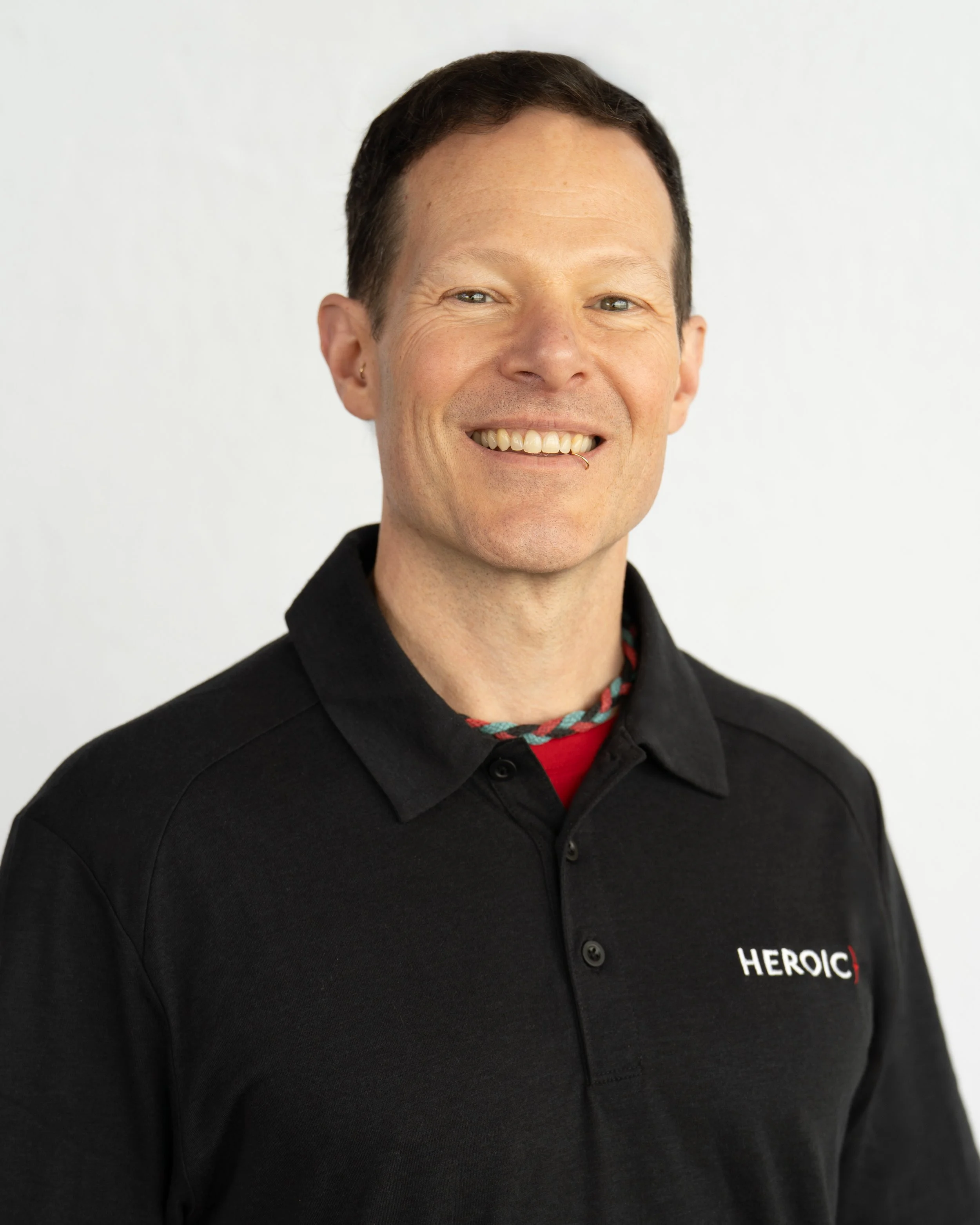 Close-up of a smiling man with short dark hair, wearing a black polo shirt with the word 'HEROIC' embroidered on it, and a colorful beaded necklace, standing against a plain white background.