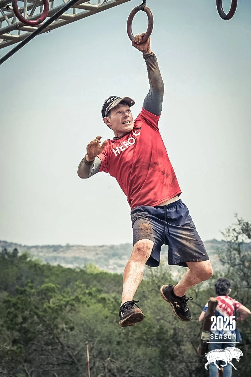 A man wearing a red T-shirt with the word 'HERO' on it and a black cap is participating in a Tough Mudder obstacle course. He is hanging from gymnastic rings suspended from a metal structure, with a dirt-covered face and mud-stained legs. The background shows trees and a cloudy sky. The image includes text indicating it's the 2025 season.