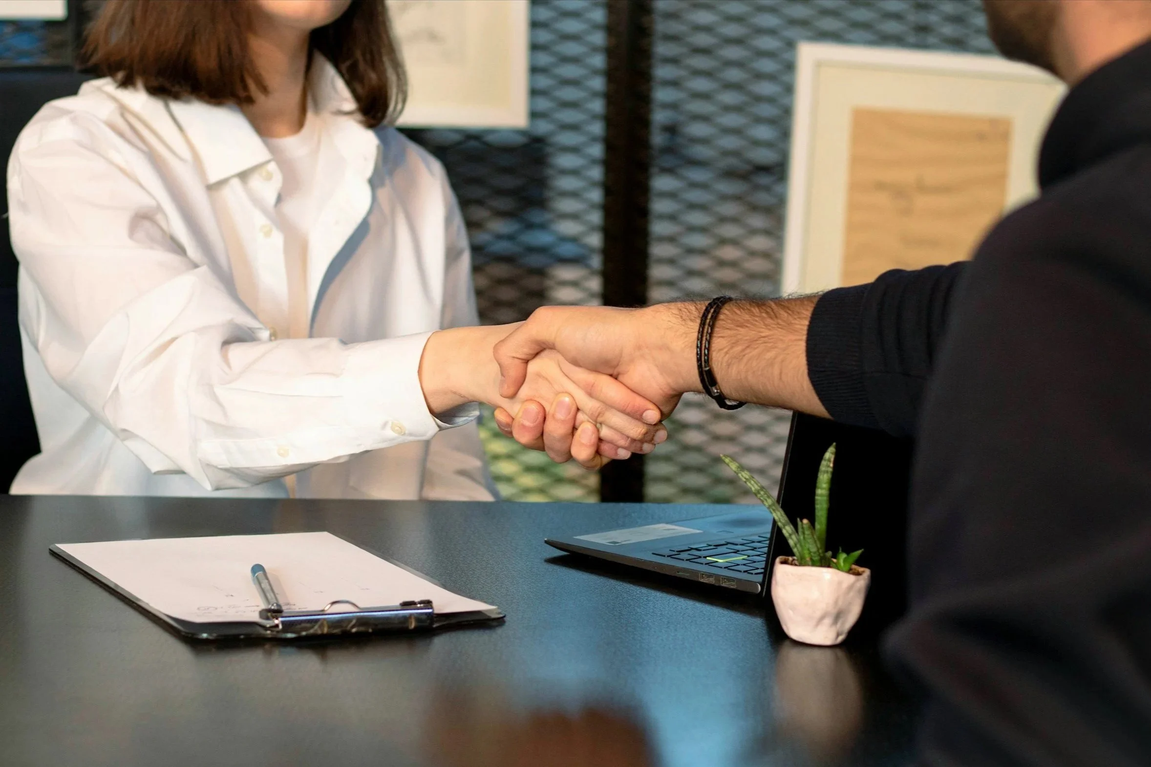 Two people shaking hands across a table with a notebook, pen, laptop, and small potted plant.