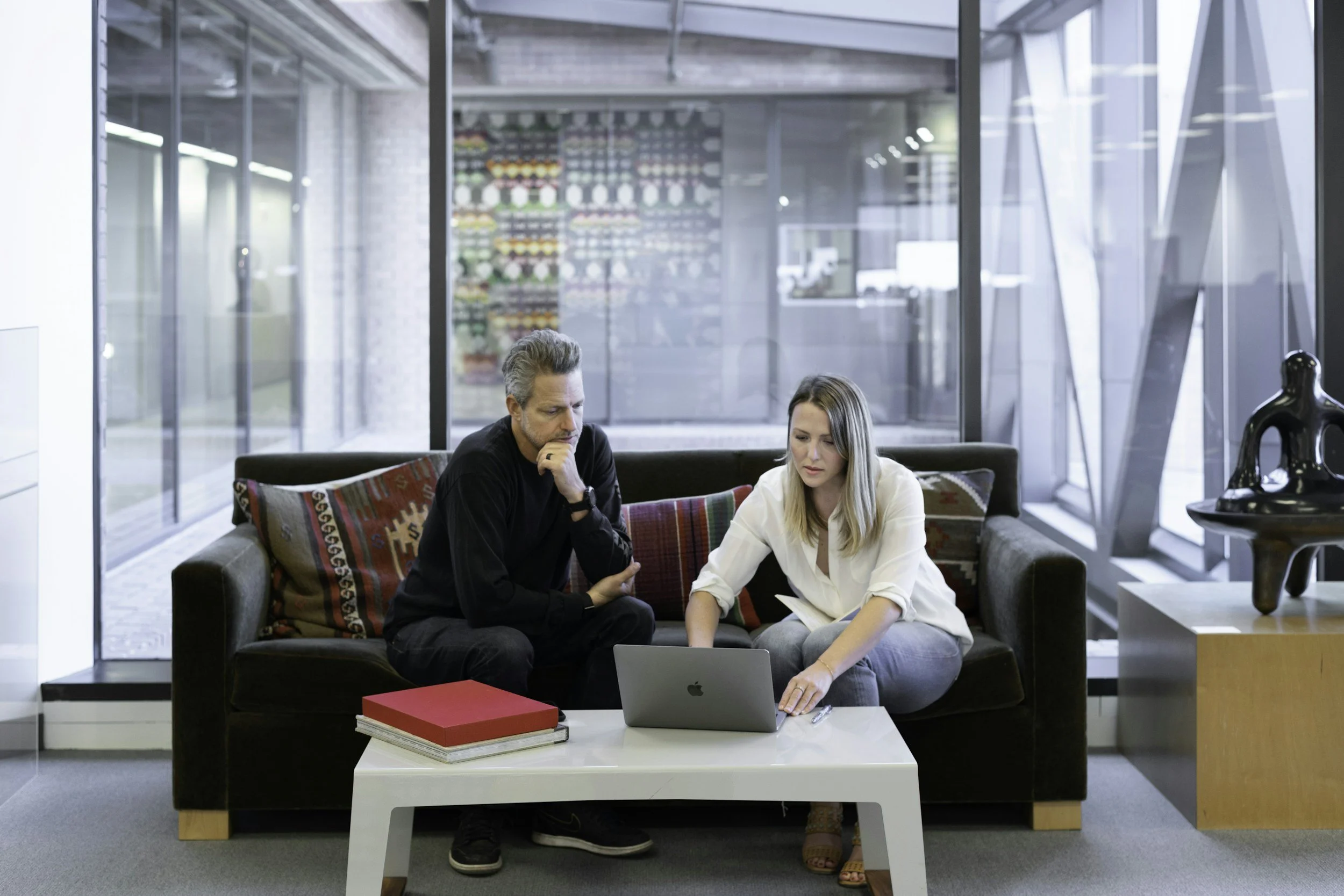 Two people sitting on a sofa in a modern office lobby, looking at a laptop, with books and a red box on a white coffee table in front of them.