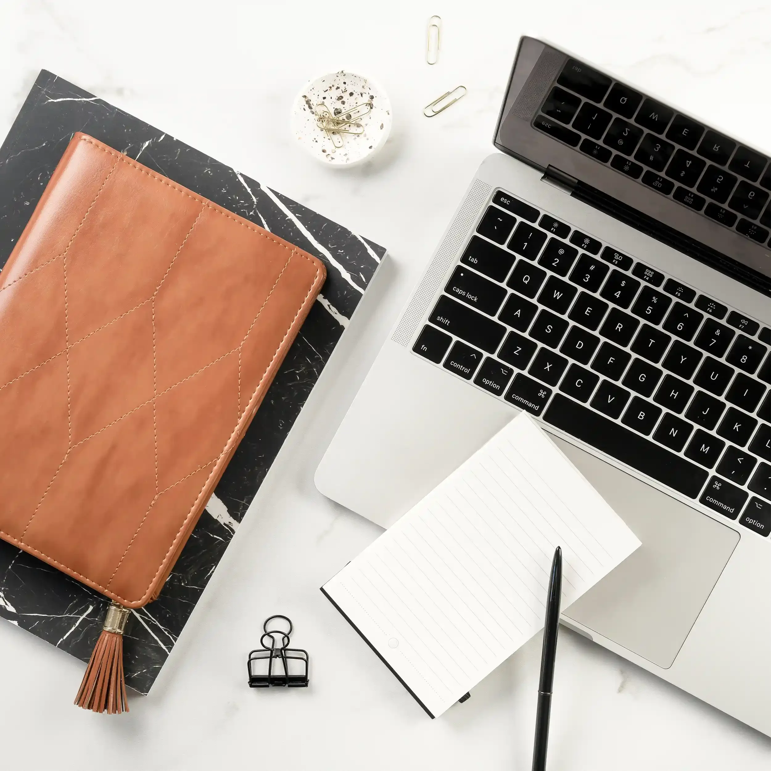 Overhead view of a workspace with a silver laptop, a brown notebook, a black marble notebook, a white tablet with a stylus, a small white bowl with gold paper clips, a few paper clips scattered, a black binder clip, a pair of black scissors, and a gold tassel keychain on a white surface.